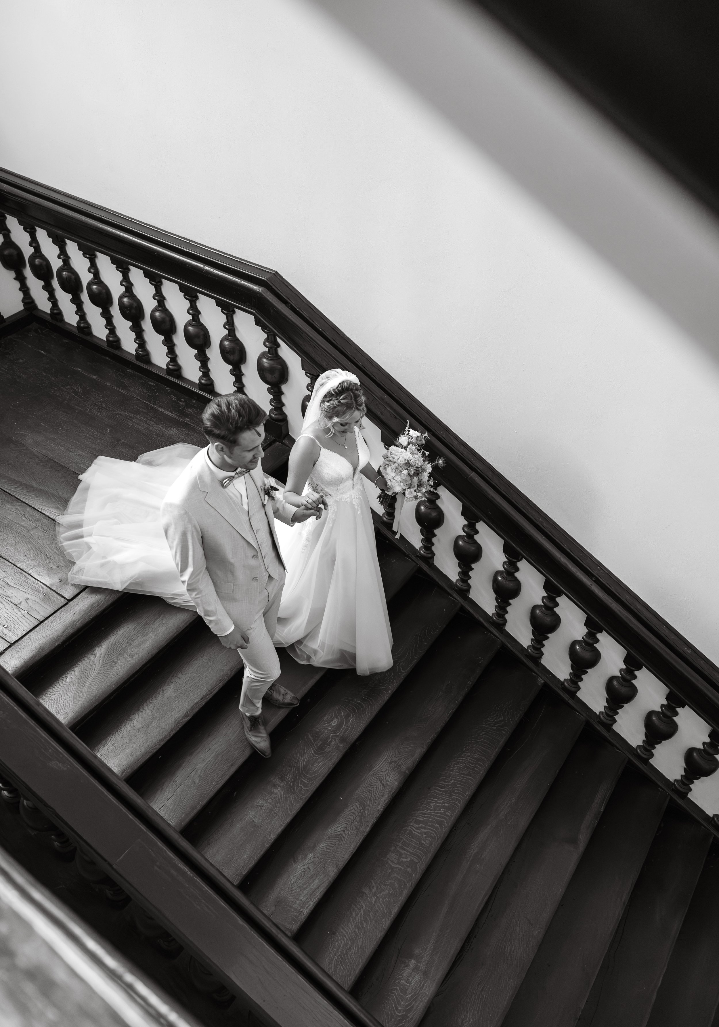 Schwarz-weiß-Foto eines Brautpaares, das eine Treppe in einem alten Gebäude hinuntergeht, die Braut hält einen Blumenstrauß.