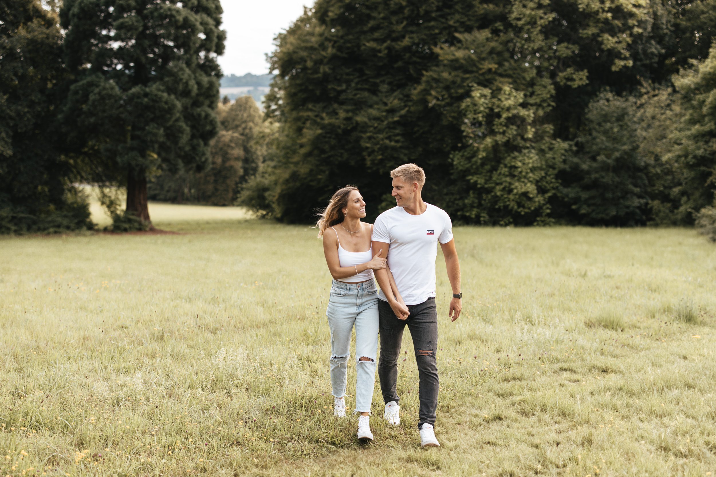 Paar läuft Hand in Hand durch eine grüne Wiese in einem Park mit Bäumen im Hintergrund.