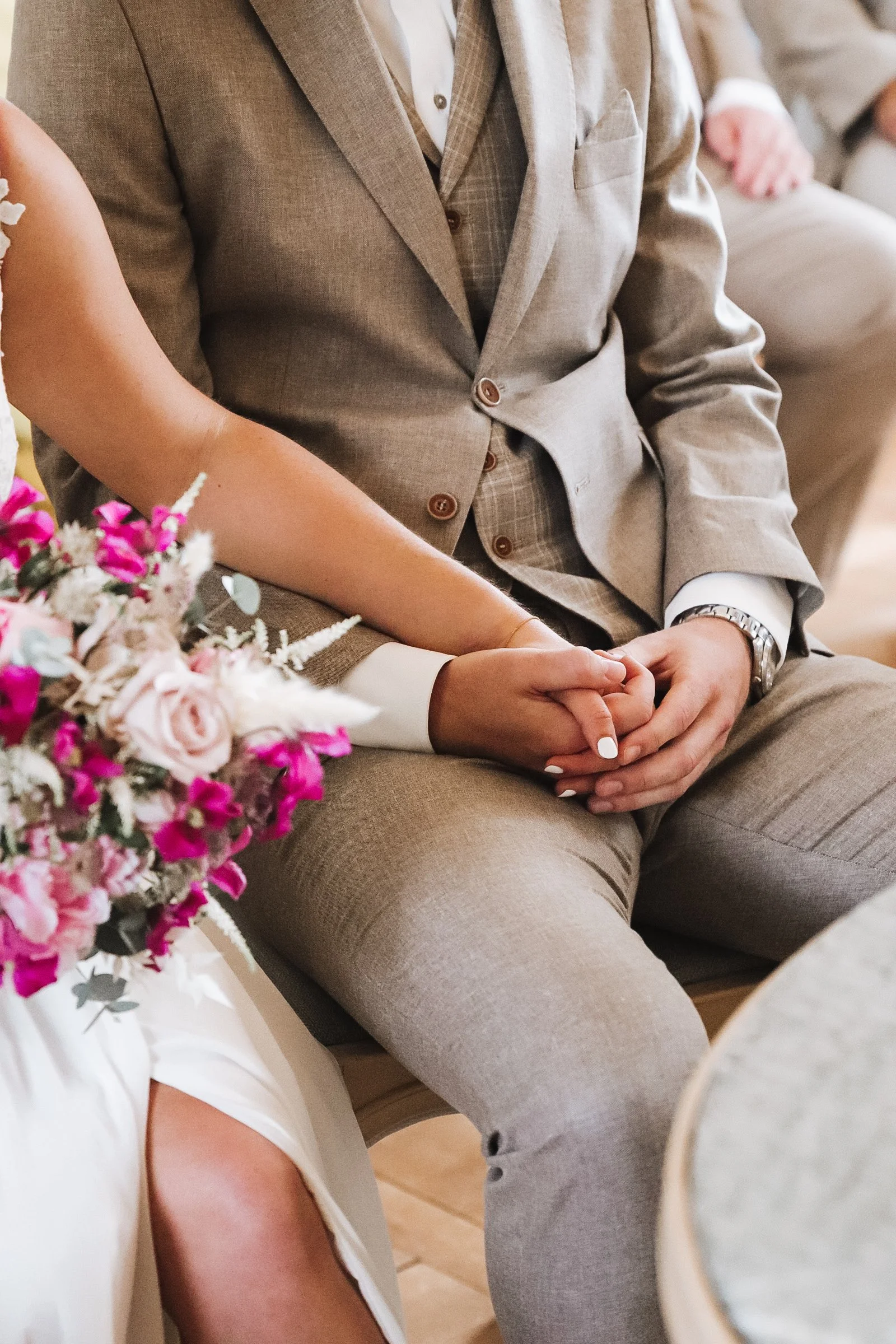 Ein Brautpaar sitzt während einer Hochzeit, hält Händchen, die Braut trägt ein weißes Kleid und hält einen Blumenstrauß, der Bräutigam trägt einen beigen Anzug.