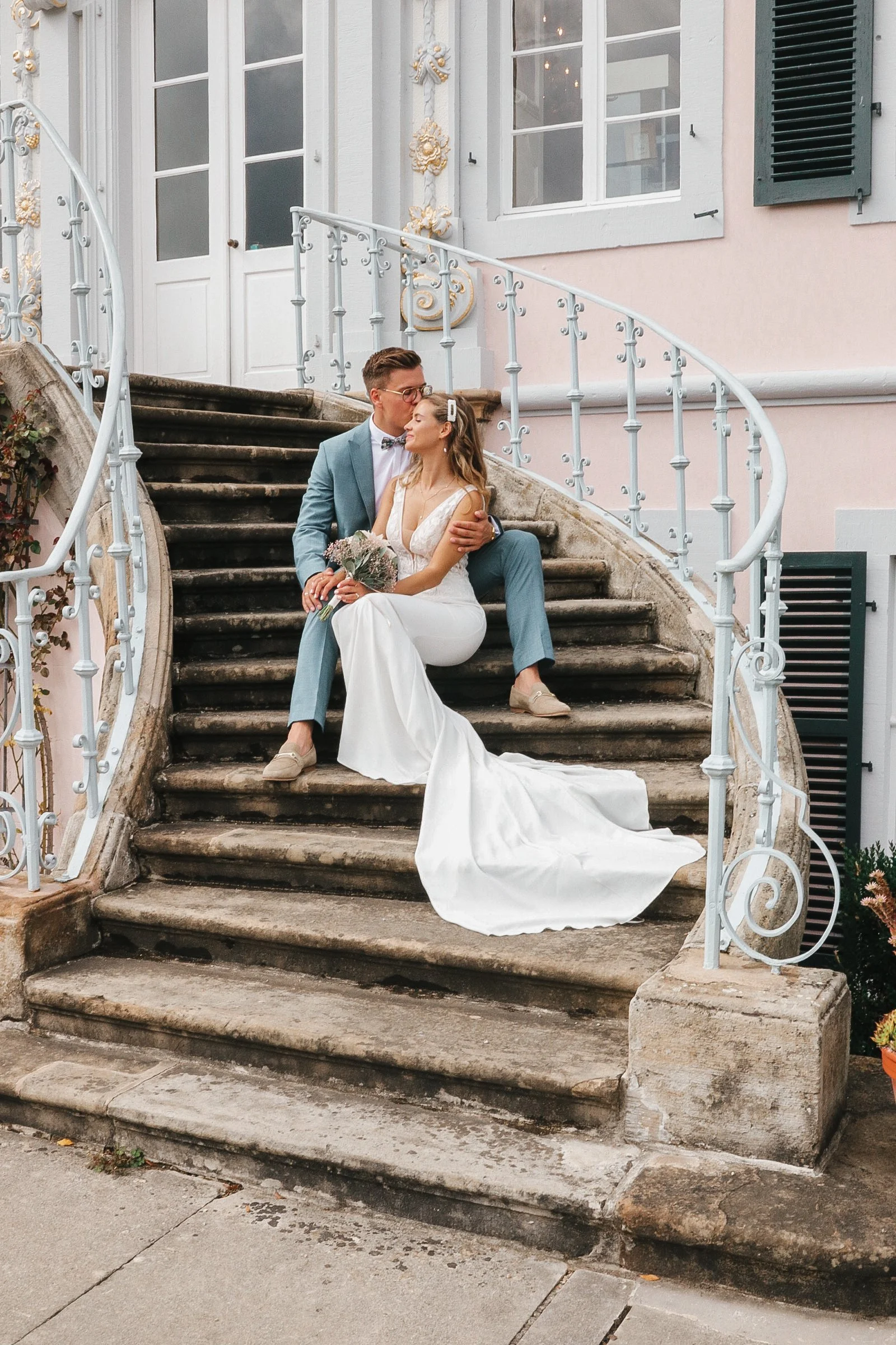 Ein Brautpaar sitzt auf einer Treppe vor einem Haus. Der Bräutigam trägt einen blauen Anzug und die Braut ein weißes Hochzeitskleid. Er küsst sie auf die Stirn und sie hält einen Blumenstrauß.