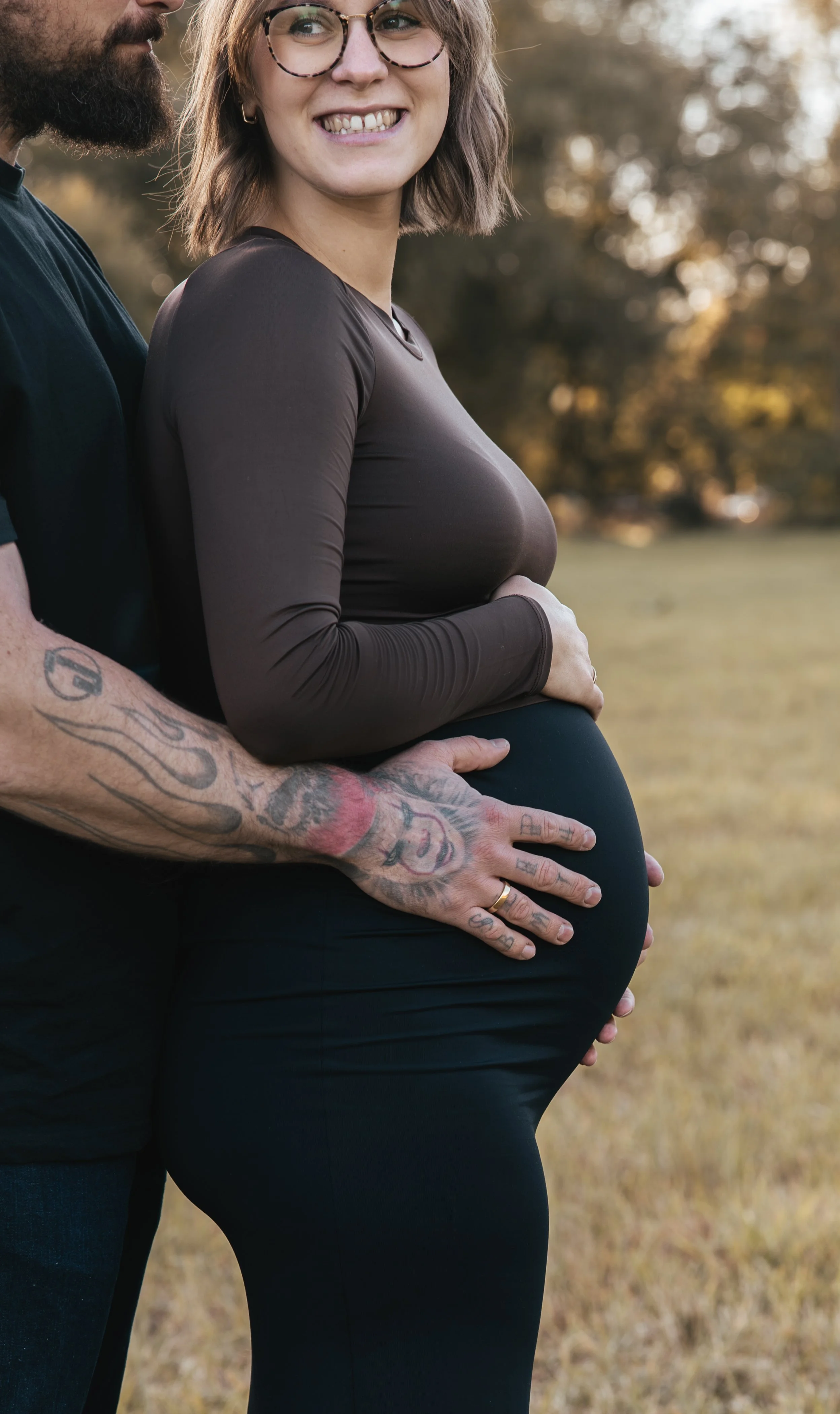 Ein schwangerer Frau, die von einem Mann umarmt wird, der Tattoos an den Armen hat, im Freien bei Sonnenuntergang.