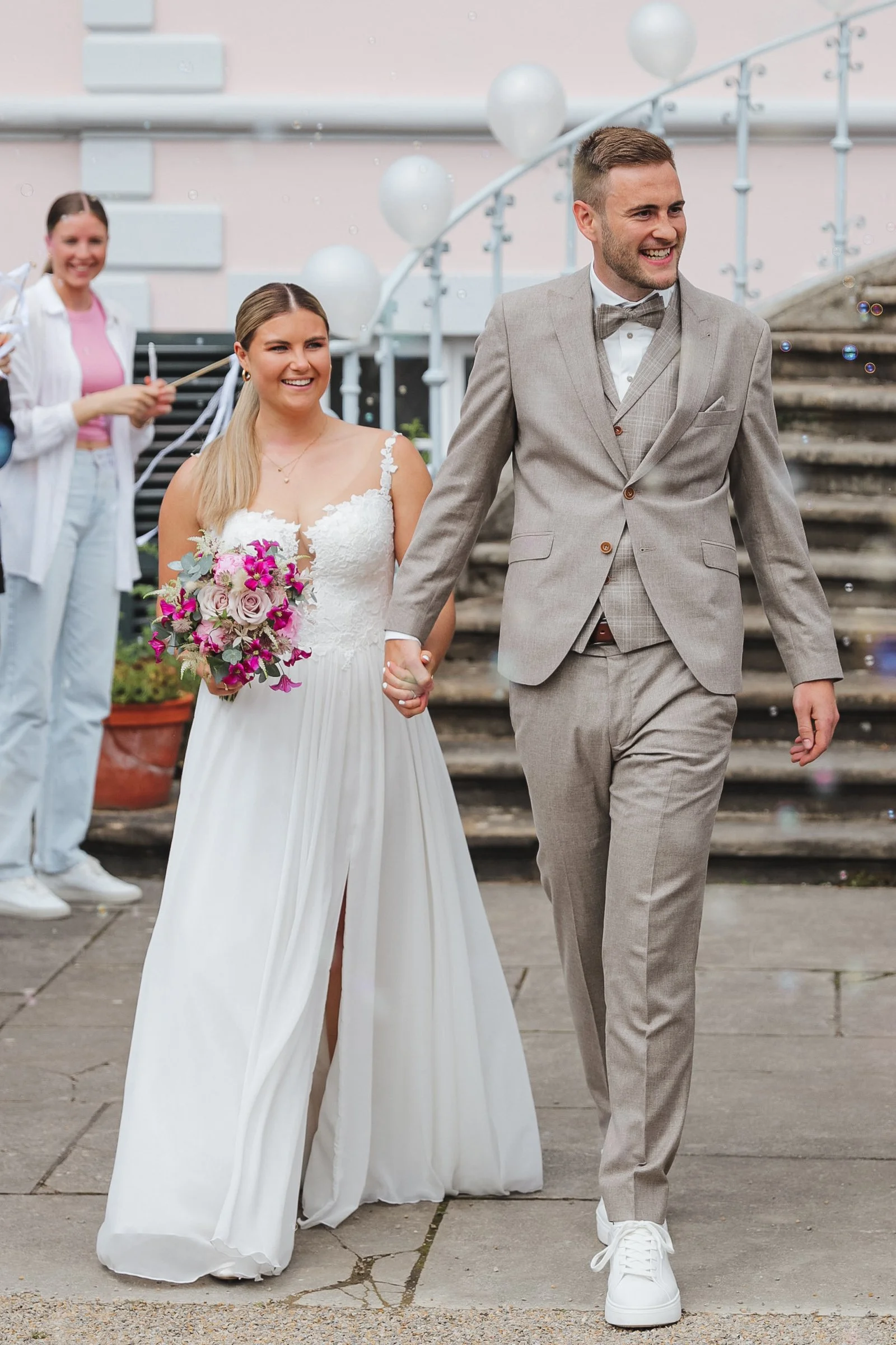 Ein frisch verheiratetes Paar kommt Hand in Hand aus dem Standesamt. Die Braut trägt ein weißes Kleid und hält einen Blumenstrauß, während der Bräutigam einen beigen Anzug mit Fliege trägt. Im Hintergrund feiern Gäste, die Seifenblasen blasen.