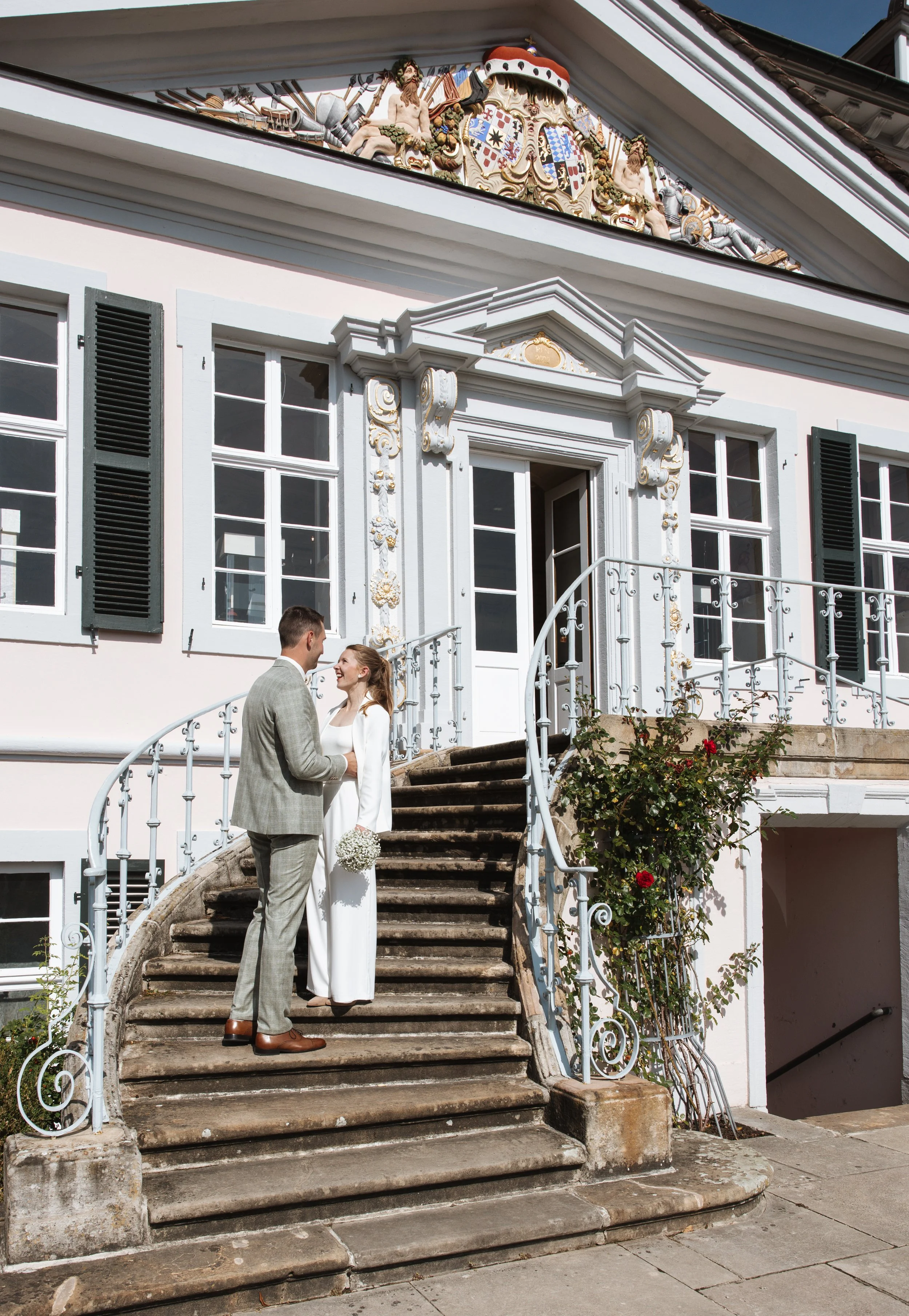 Ein verliebtes Paar steht auf einer alten Steintreppe vor einem historischen, weiß gestrichenen Gebäude mit kunstvollen Fassadendetails und dunkelgrünen Fensterläden. Der Himmel ist klar und blau.