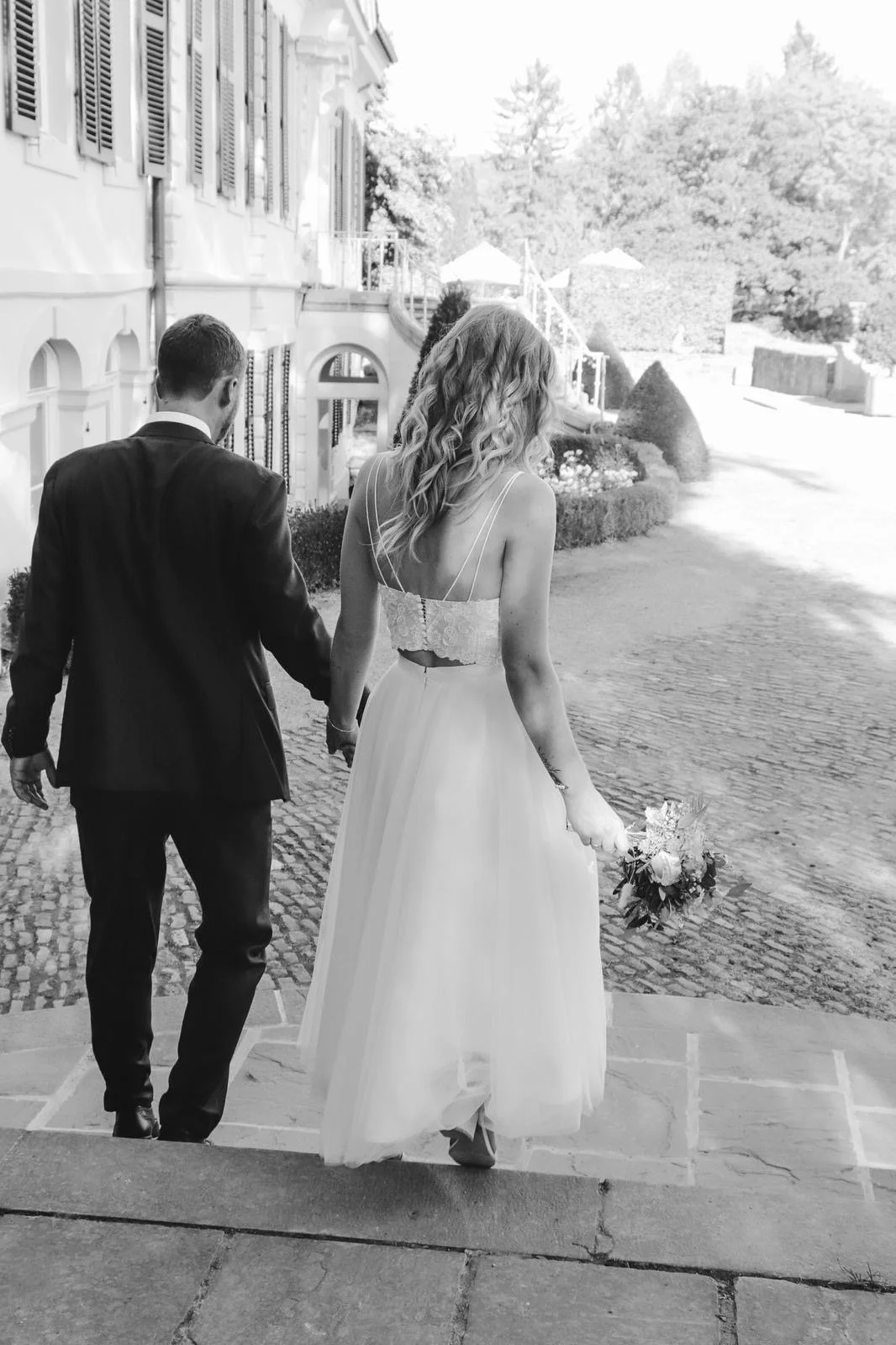 Braut und Bräutigam gehen Hand in Hand auf einer steinernen Treppe bei Sonnenlicht, im Freien, vor einem historischen Gebäude mit Fensterläden, umgeben von Bäumen und Gartenzierde, während sie an ihrem Hochzeitstag unterwegs sind.