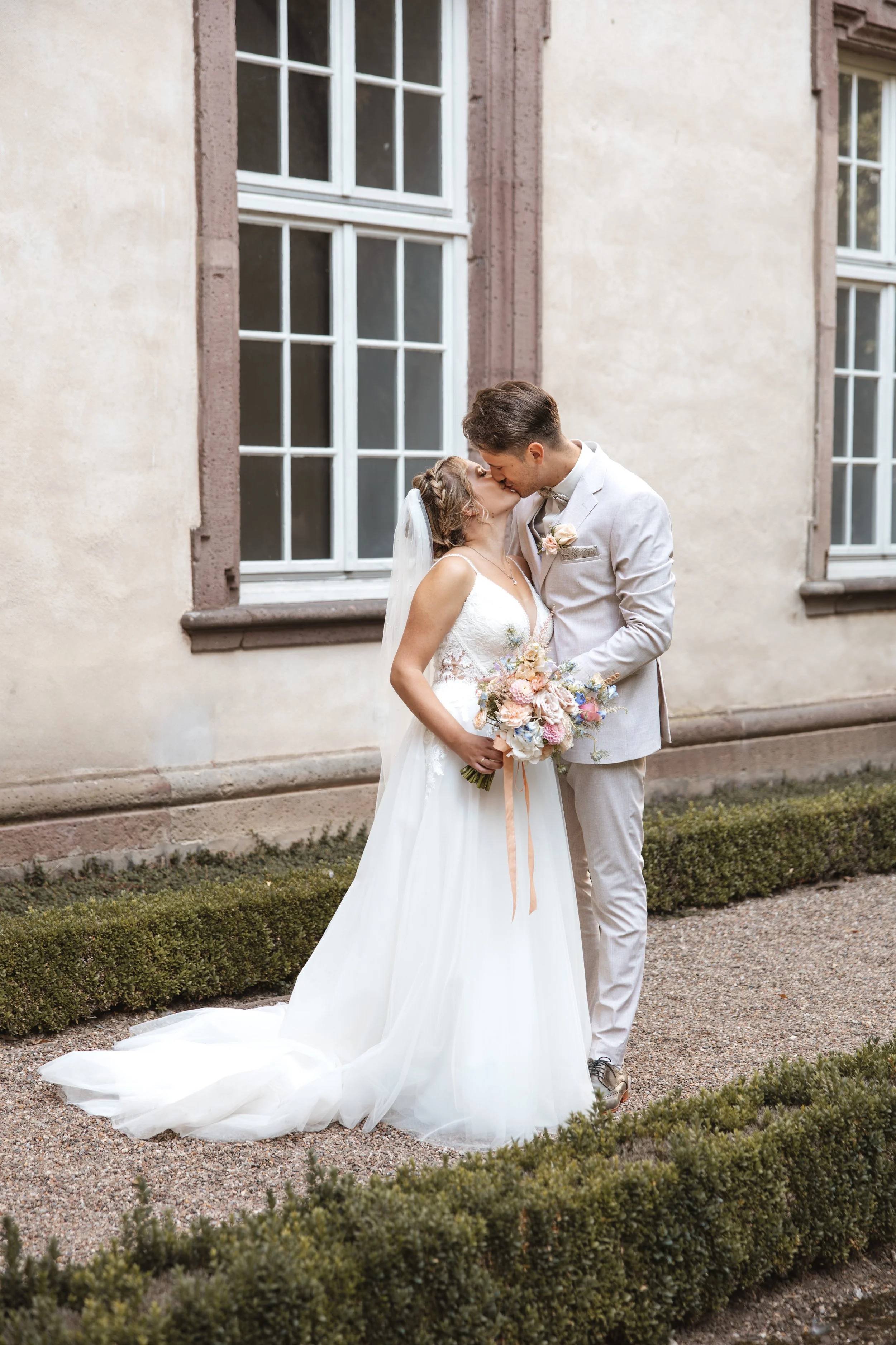 Ein frisch vermähltes Paar bei ihrer Hochzeitszeremonie, umarmend und küsst sich vor einem alten Gebäude mit großen Fenstern.