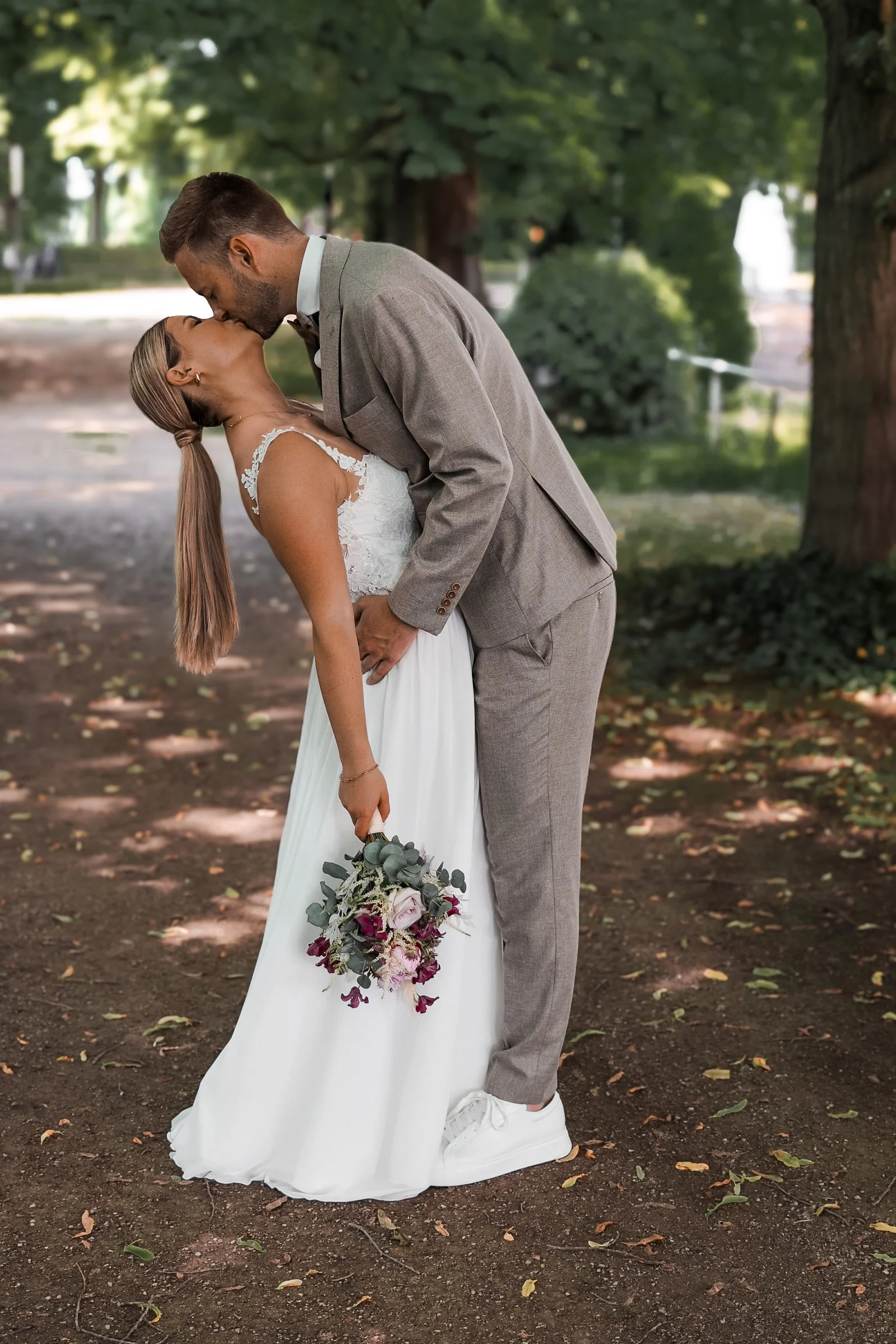 Ein frisch verheiratetes Paar, das sich küsst, steht in einem Park. Der Mann trägt einen beigenAnzug, die Frau ein weißes Brautkleid und hält einen Brautstrauß in der Hand.