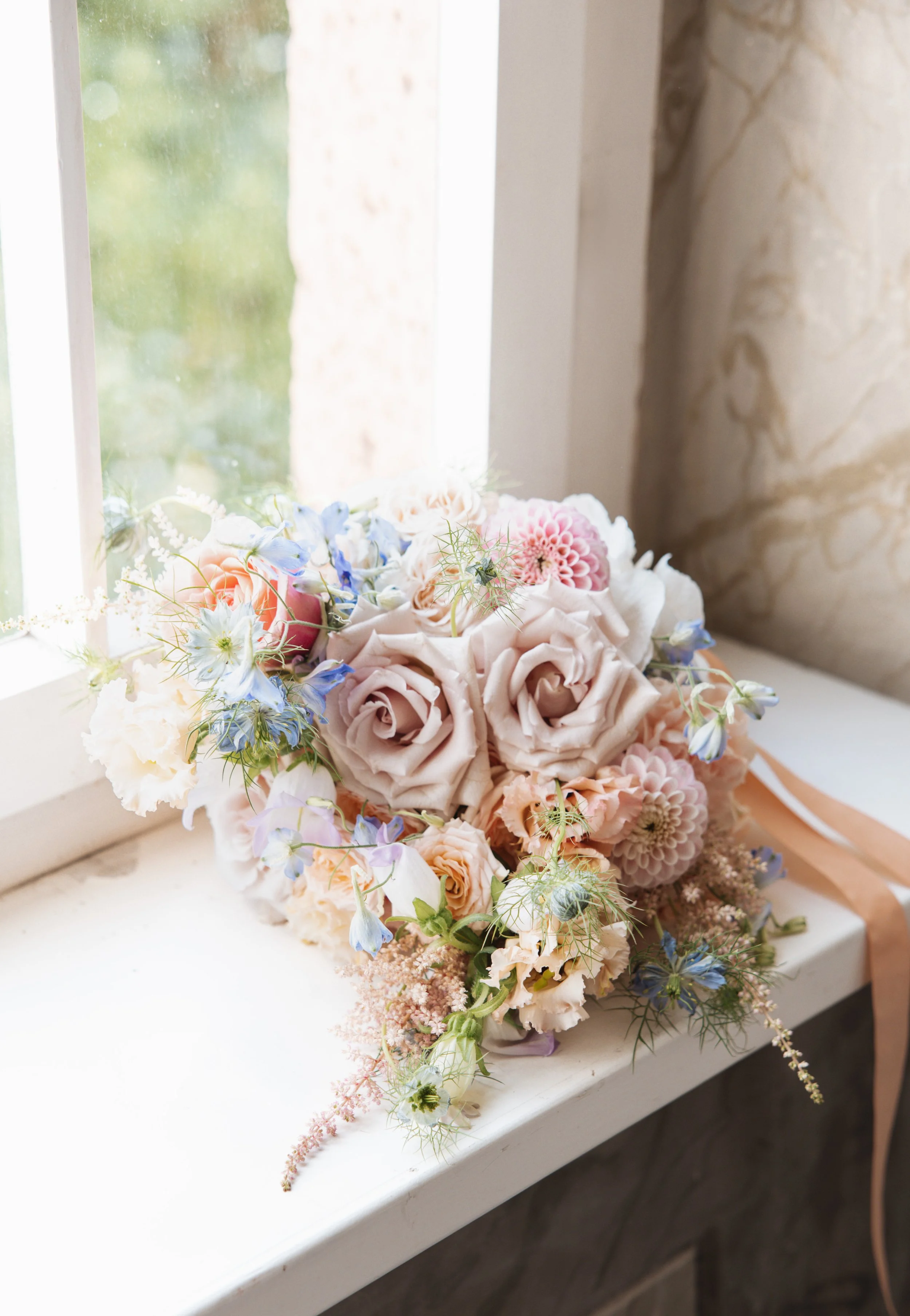 Ein Blumenstrauß mit rosa, blauen und weißen Blumen auf einer weißen Fensterbank neben einem Fenster und einer Steinwand.