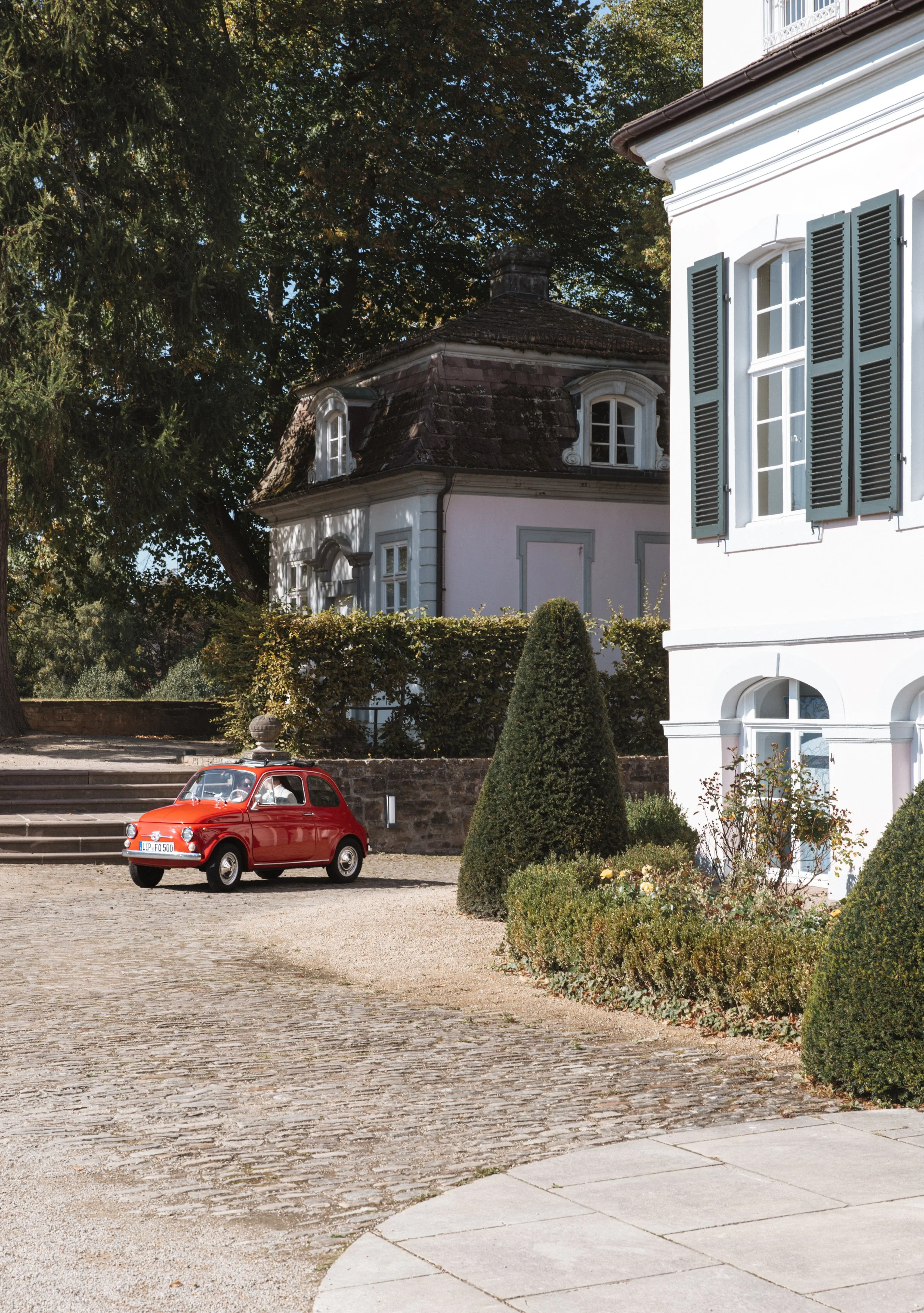 Ein rotes Oldtimer-Auto steht vor einem historischen Gebäude mit dunkelgrünen Fensterläden, umgeben von gepflegten Büschen und Bäumen bei sonnigem Wetter.