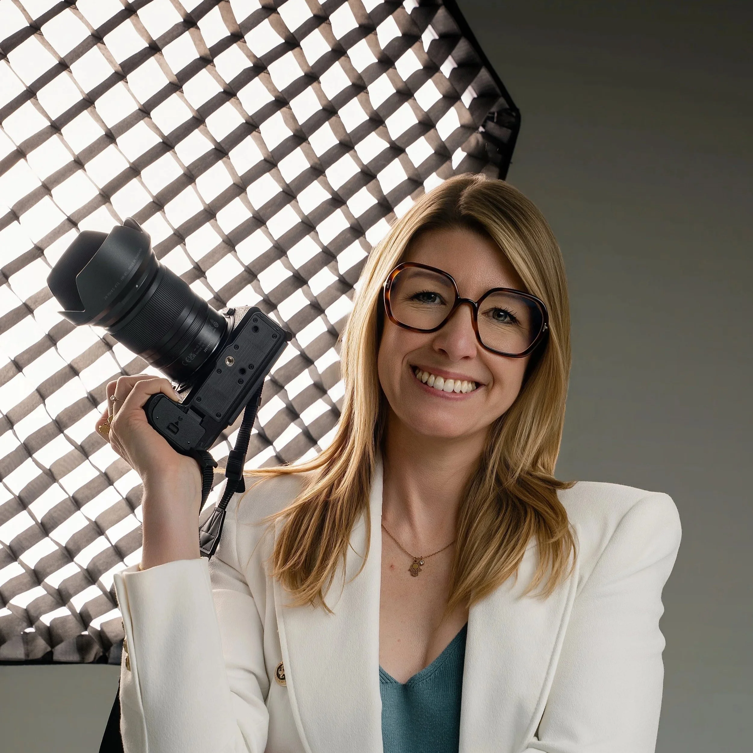 Close-up van een glimlachende vrouw met bruin haar, draagt grote tortoise-beeld schelphes en een witte blazer, onder een lichtgebobbelde dienst van een cameralens zittend.