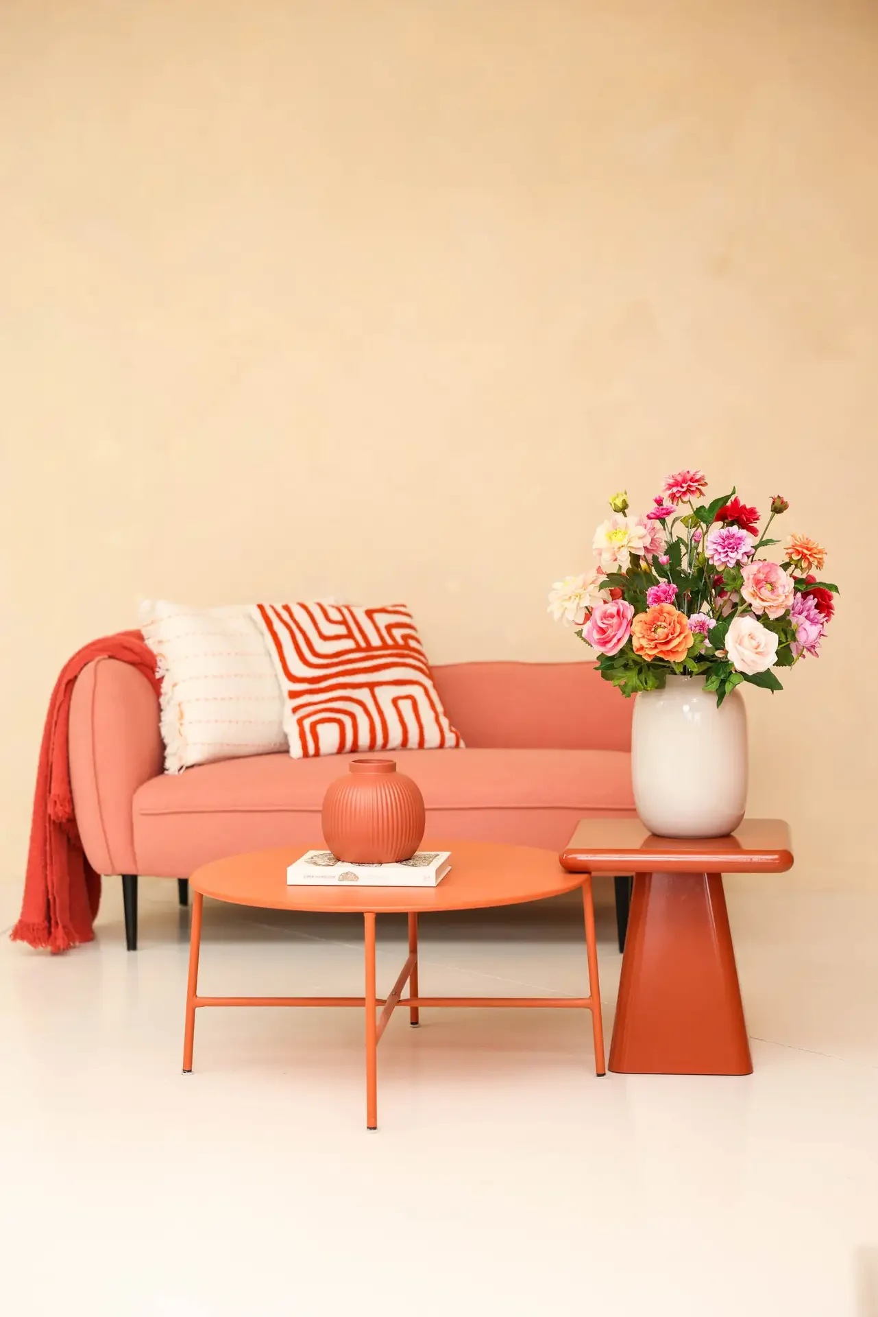 Een rosa bank met een rood-wit geblokt deken en een sierkussen, een ronde roze salontafel met een vaas met bloemen, een rechthoekige bijzettafel met een vaas met bloemen, tegen een lichtbeige muur.