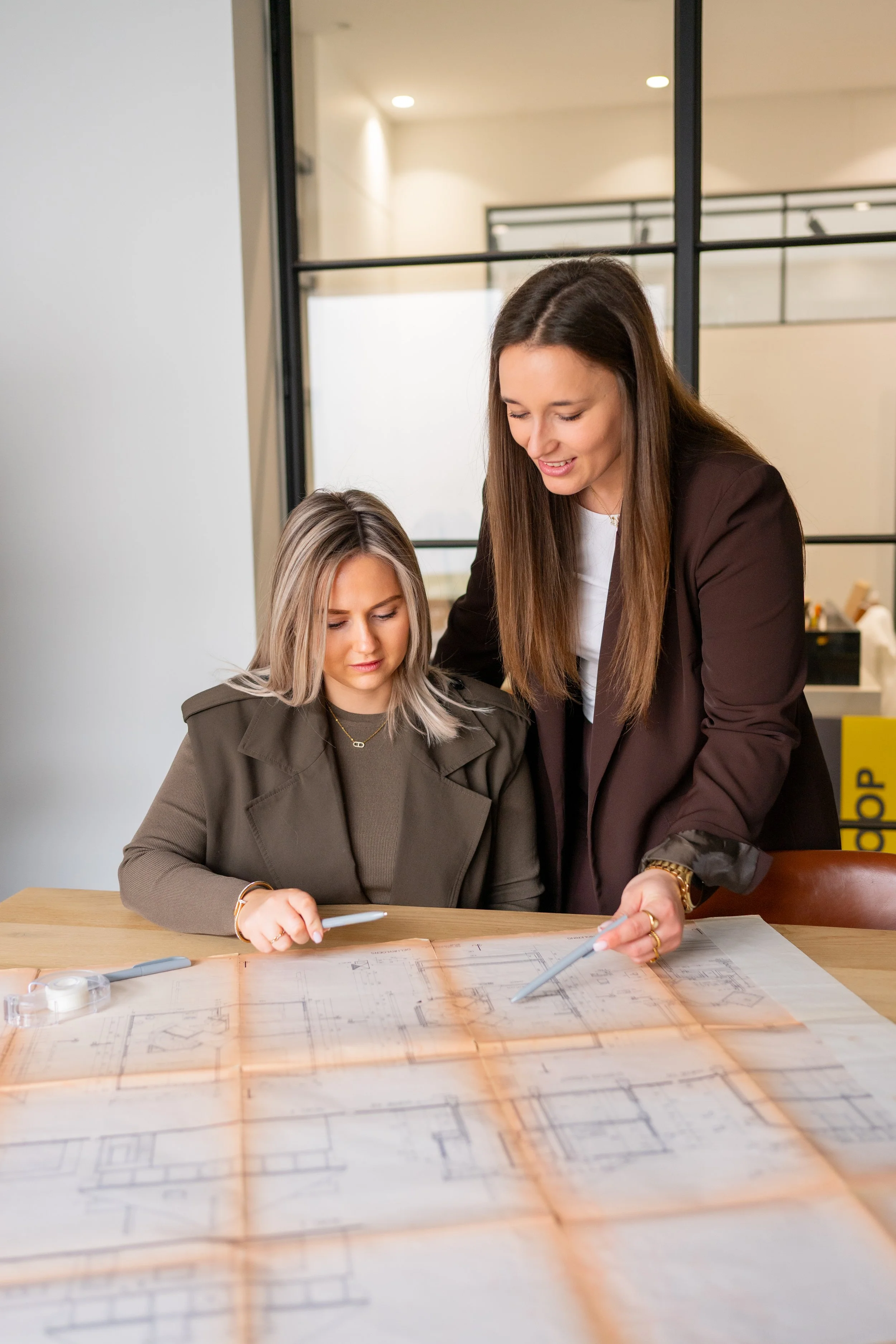 Twee vrouwen op kantoor  bespreken een bouwplan of blauwdruk op een tafel, met grote ramen op de achtergrond. beige-bruine tinten