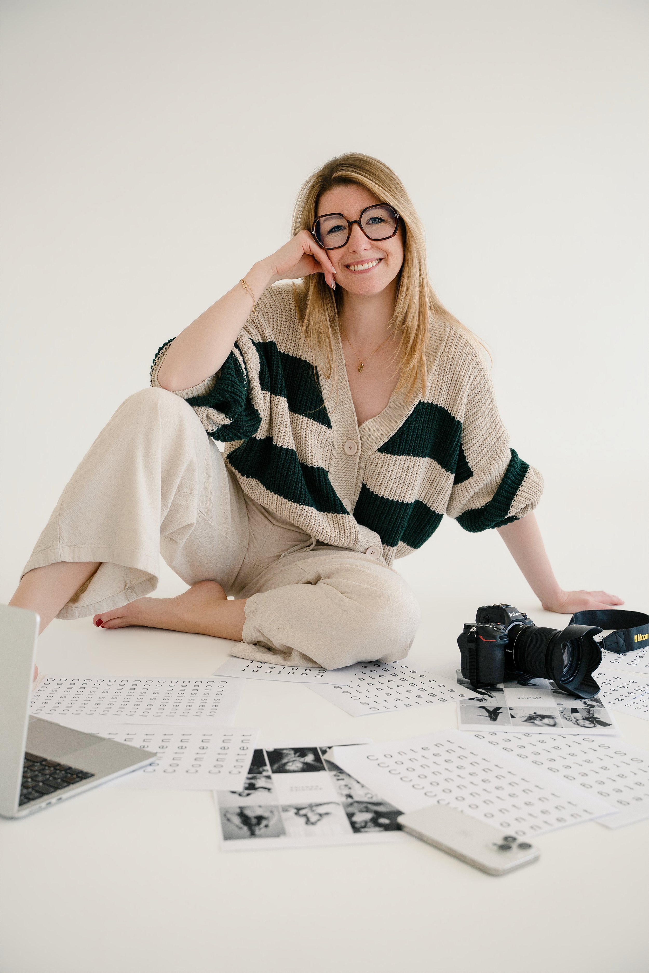 Een glimlachende fotografe met brillen zit op de vloer, omringd door fotobanden, documenten en een camera, in een witte kamer.