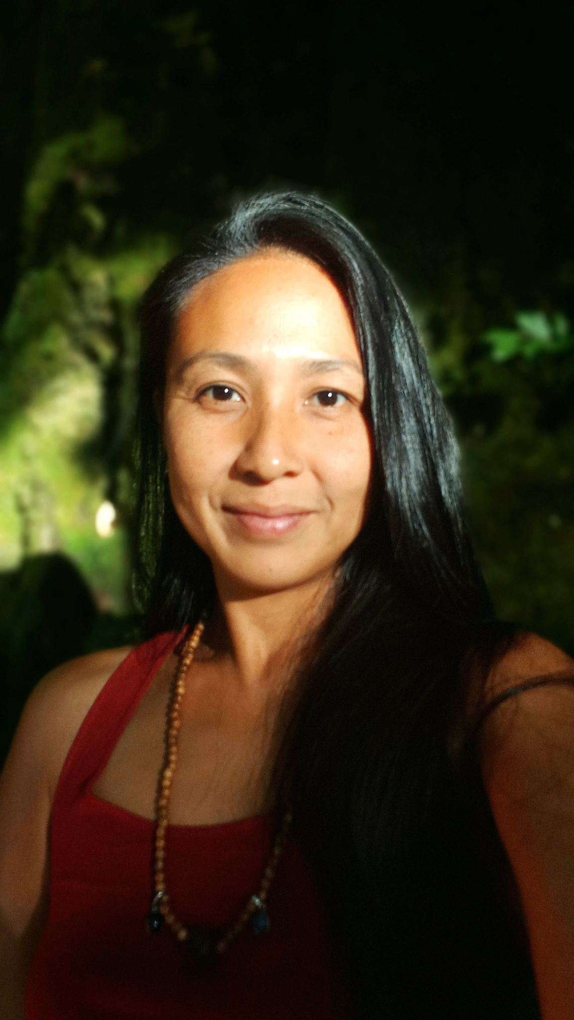 A woman taking a selfie outdoors at night, with green trees and foliage in the background.