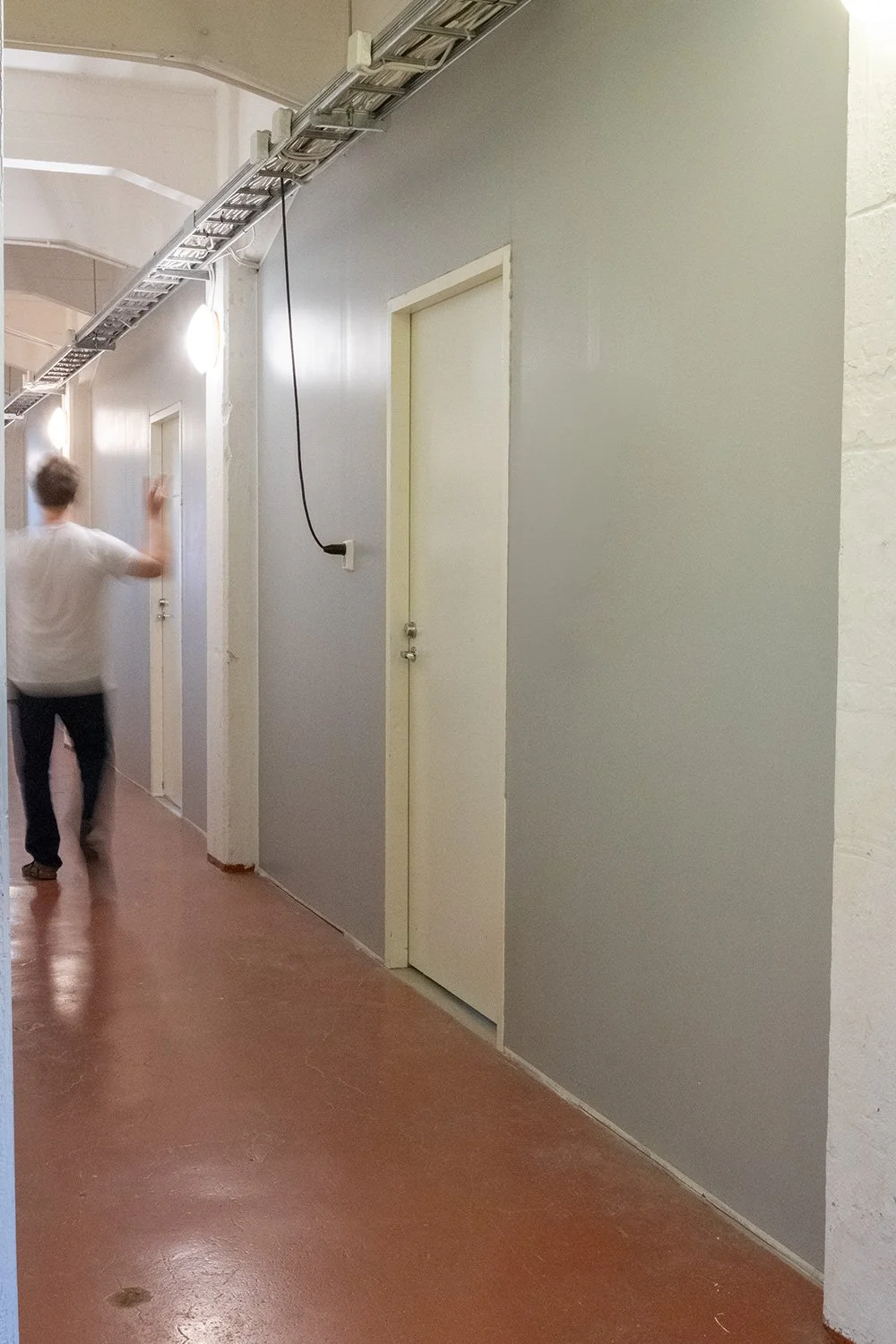 A hallway with beige walls and a wooden floor, featuring three closed white doors. An individual in a white shirt and dark pants is walking away, with their right hand raised. Overhead, there is a suspended metal cable tray with a black wire hanging from it.