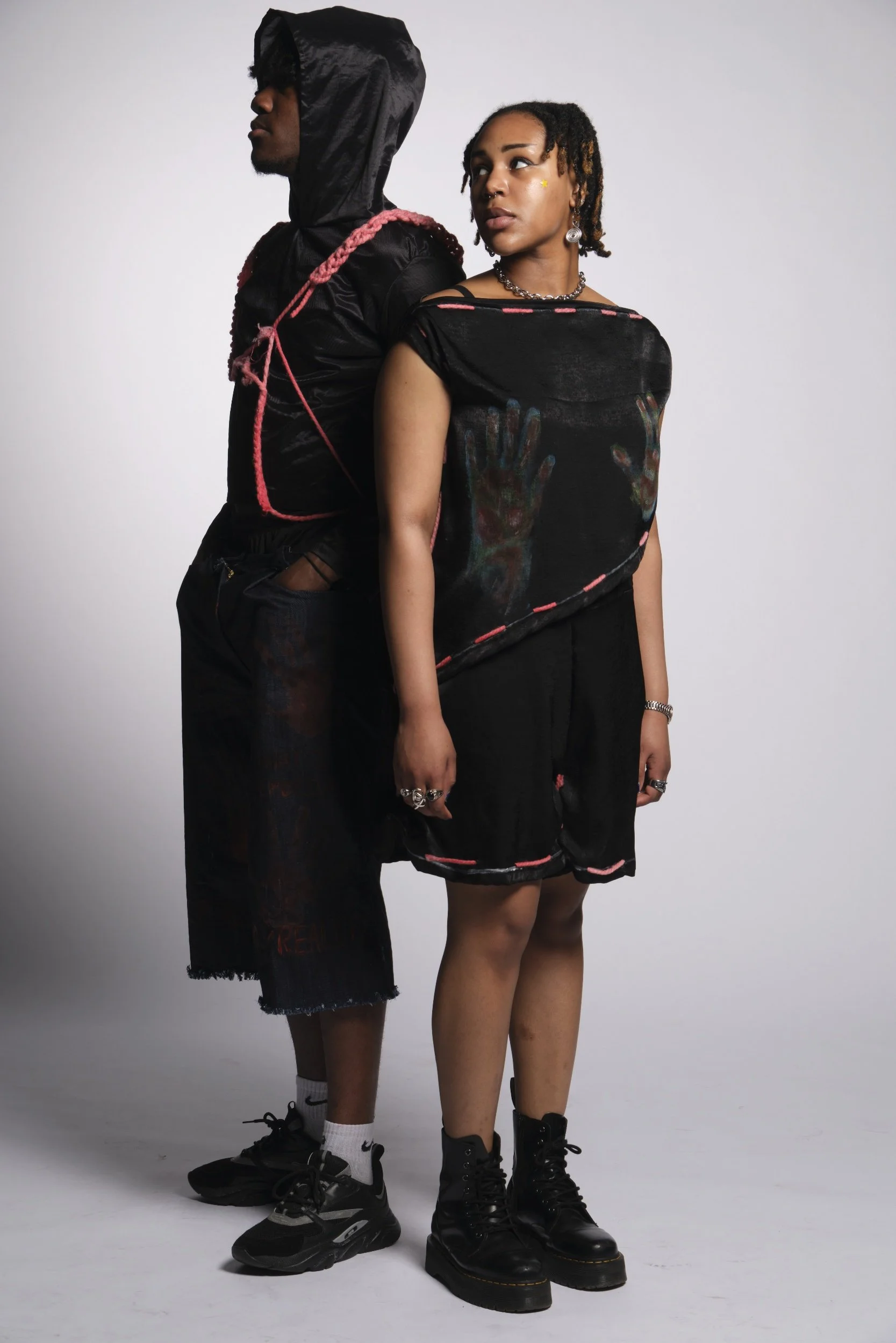 A man and woman standing back-to-back against a plain gray backdrop, dressed in dark streetwear with accessories, both looking away from the camera in a serious pose.