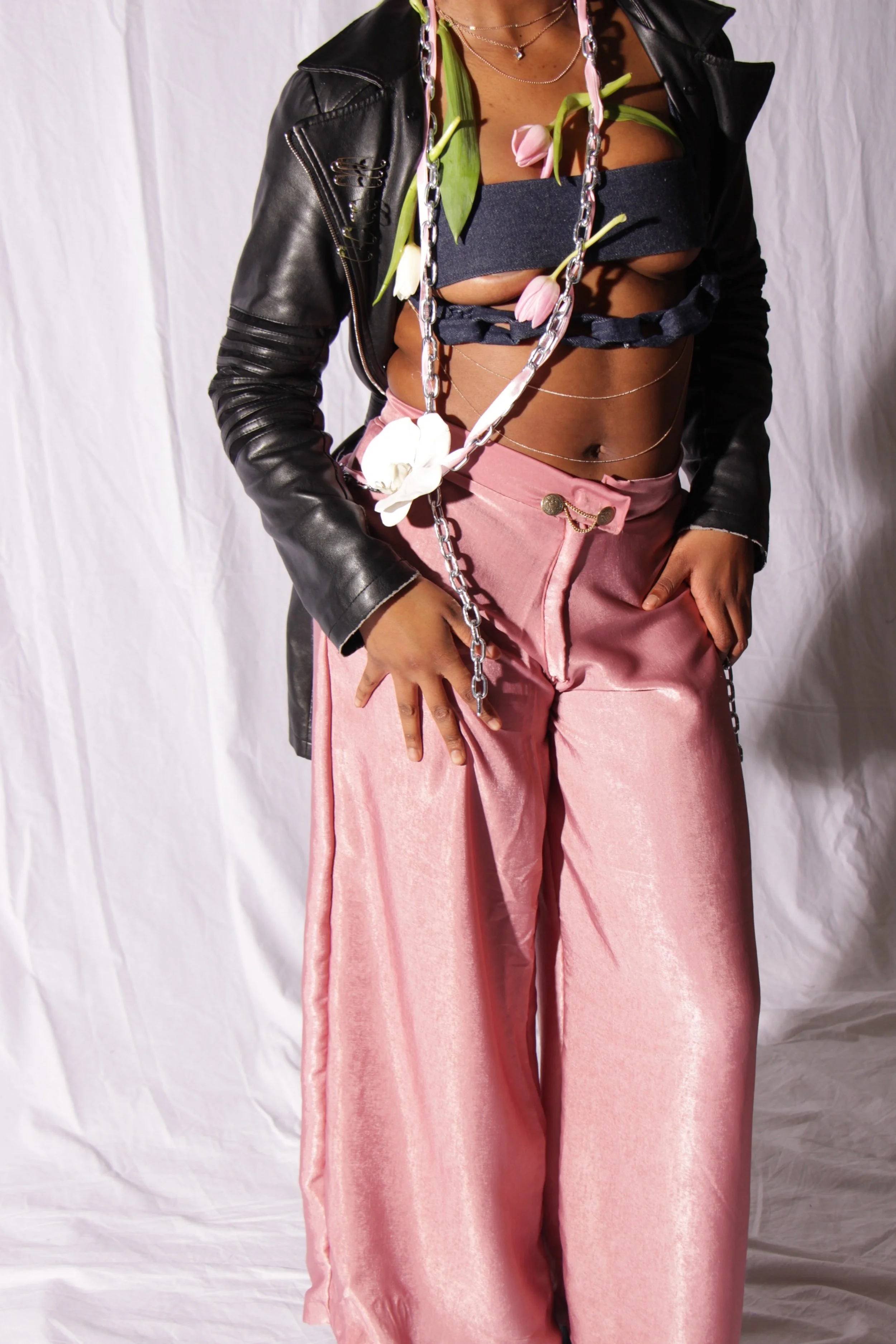 A woman wearing pink shiny wide-leg pants, a black top with cutouts, layered necklaces, and a black leather jacket. She has chains across her torso and some flowers draped around her neck and chest. The background is a plain white cloth.