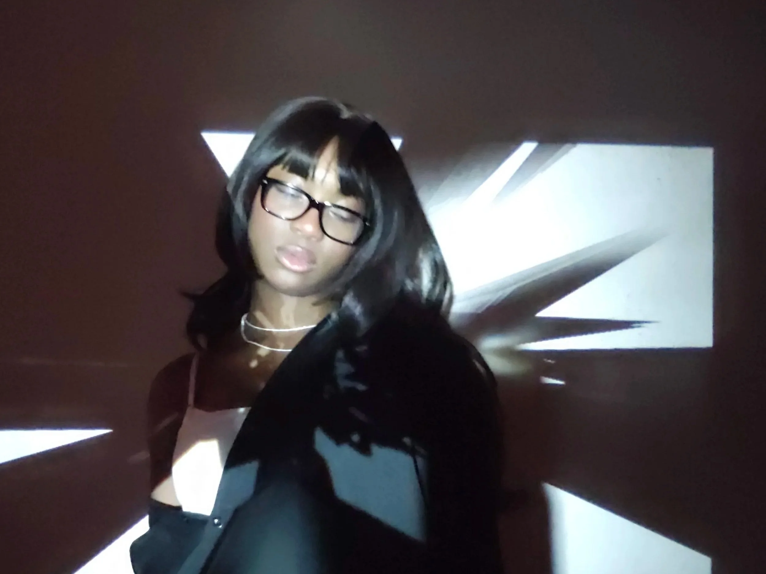 A young woman with glasses and shoulder-length black hair stands in front of a wall with abstract white light projections, creating geometric patterns around her.