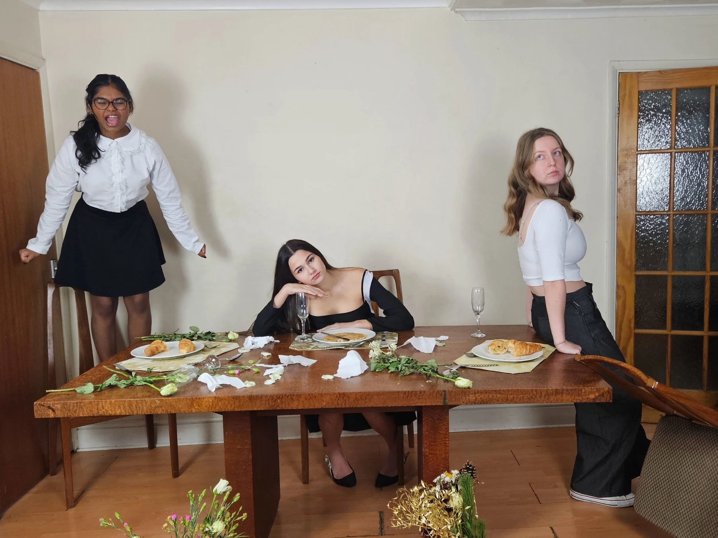 Four women at a dining table with torn paper, flowers, and food; one woman standing with eyes closed and mouth open, one sitting with her head resting on her hand, and two others with neutral expressions.