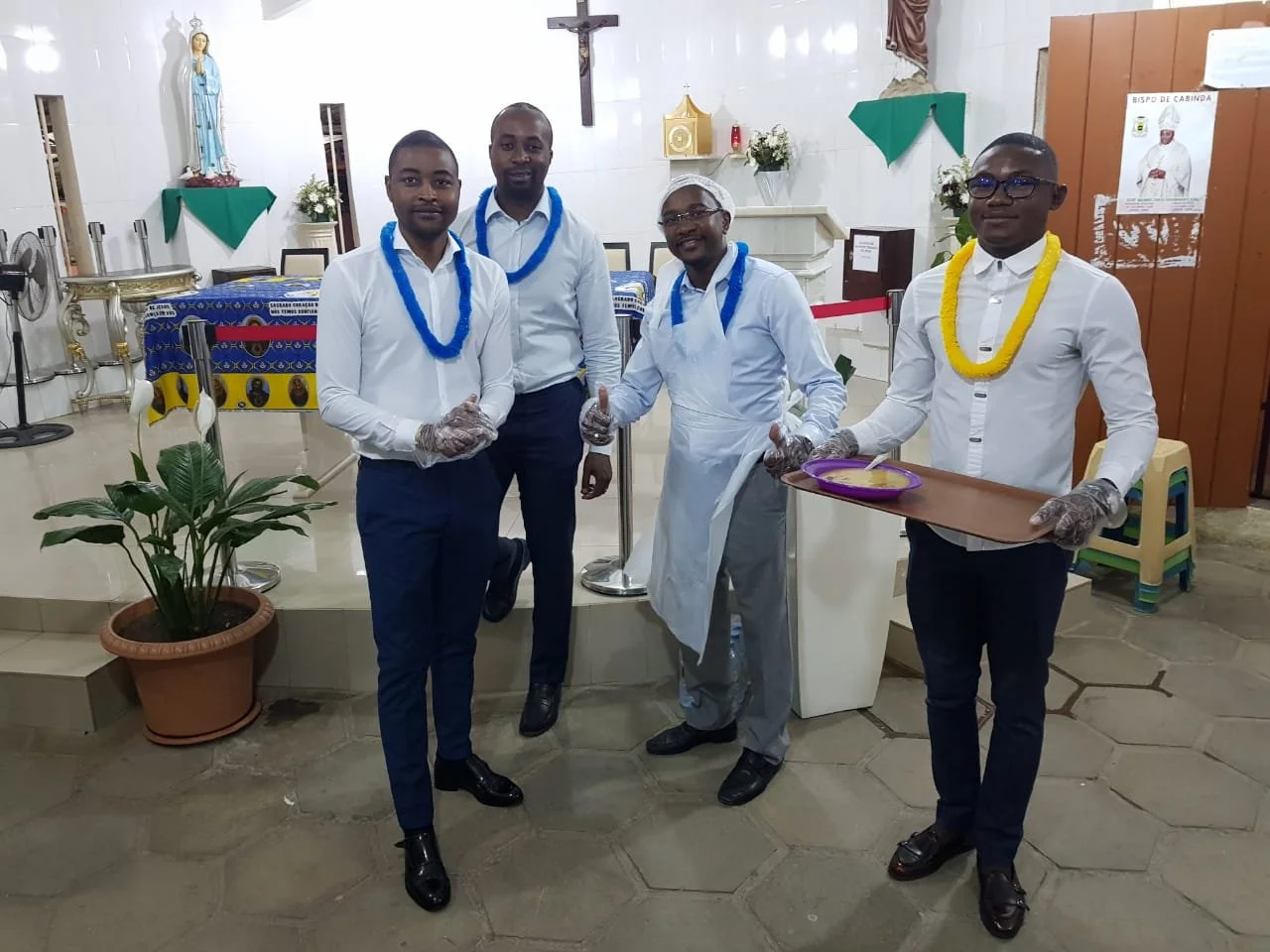Quatro homens de terno e duas mulheres vestidas de avental na igreja, participando de uma refeição comunitária, segurando pratos com comida, com uma cruz na parede ao fundo.