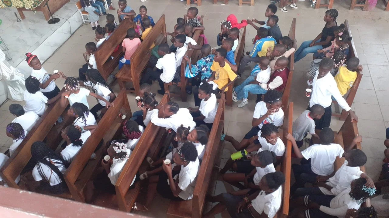 Grupo de crianças sentadas em bancos de igreja, algumas conversando e segurando copos.