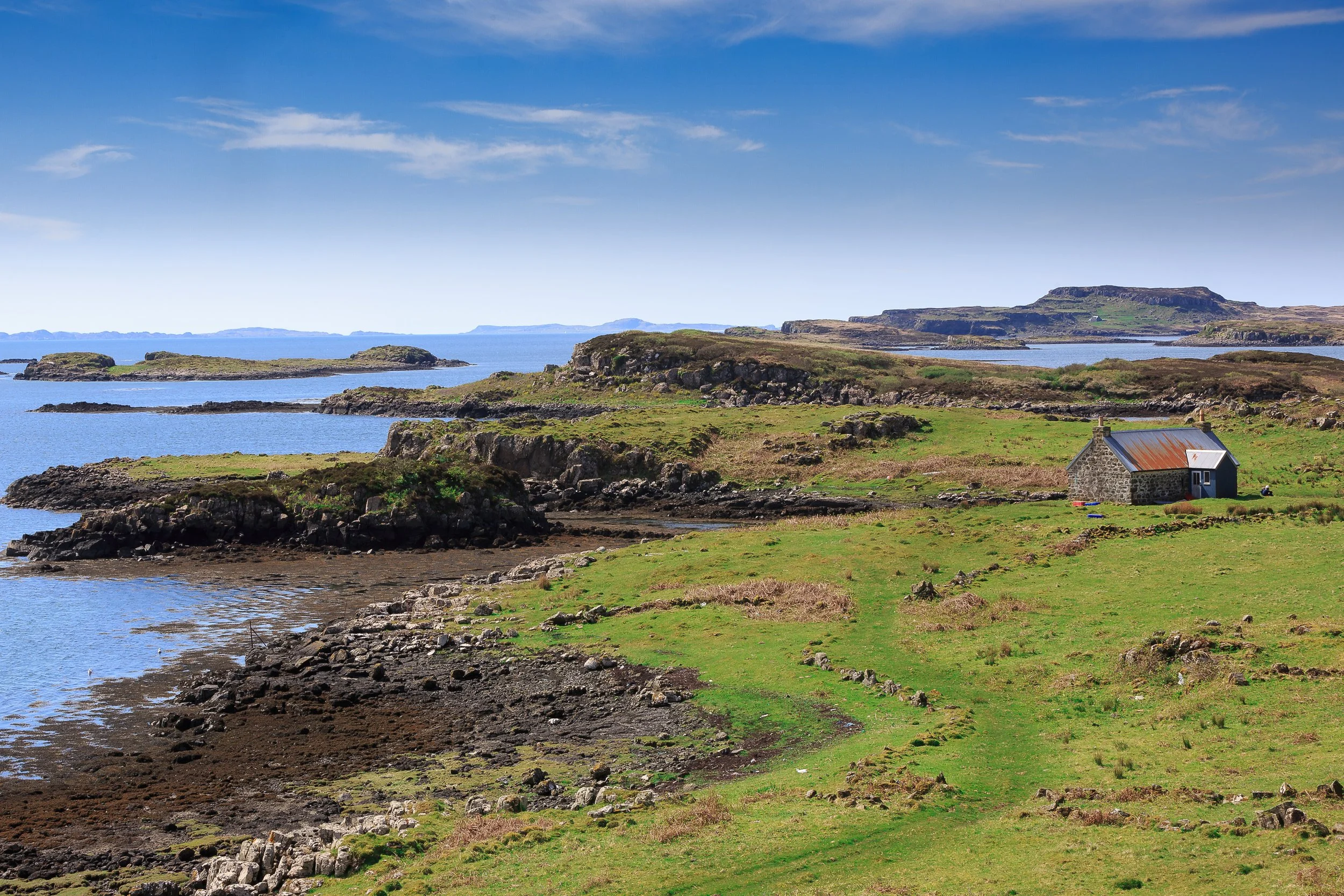 Scenic view of a coastal landscape with a small stone house, green grassy fields, rocky shoreline, and a body of water under a partly cloudy blue sky.
