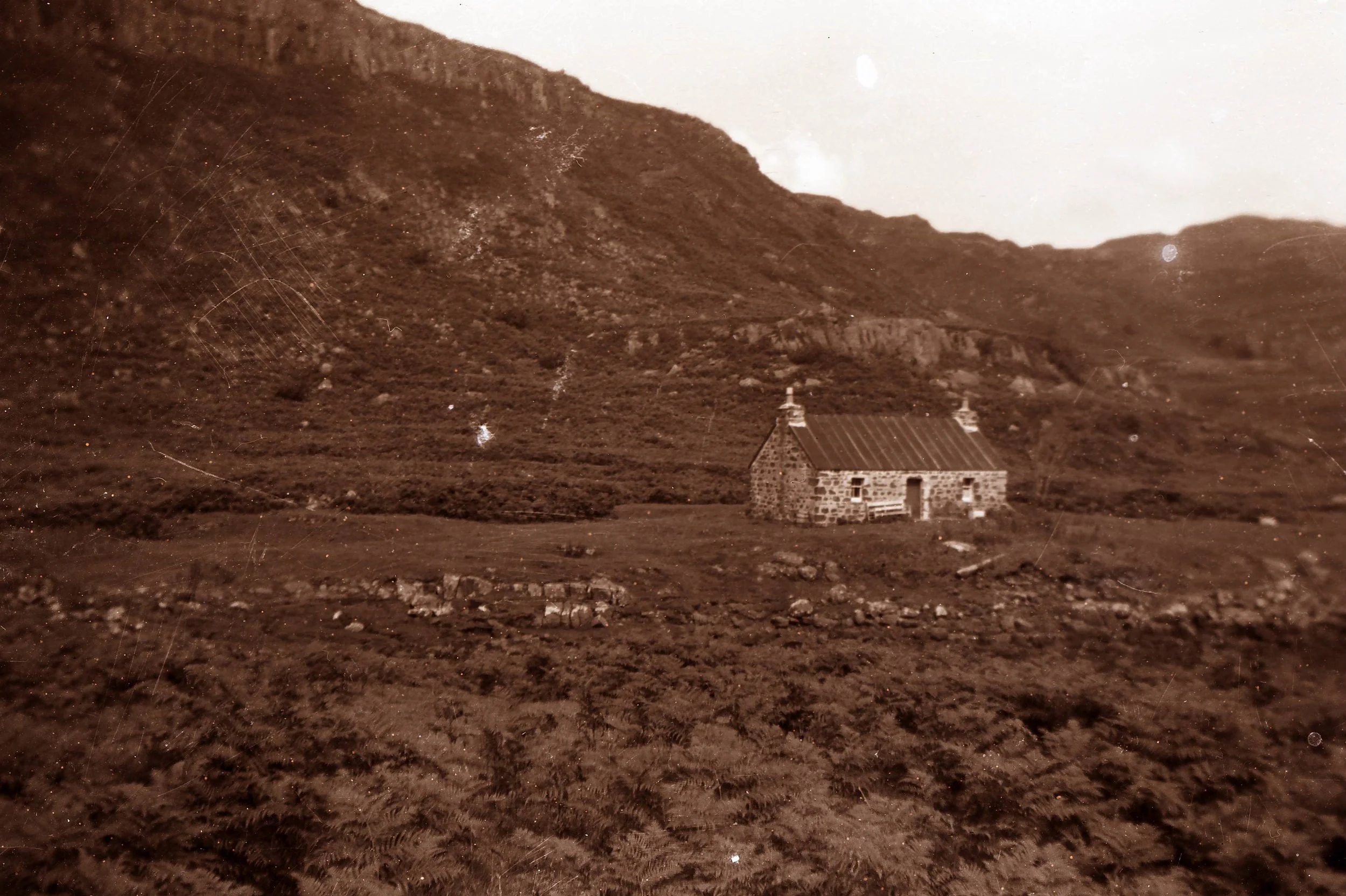 An old stone cottage with a metal roof, situated on a grassy hillside with rocky terrain, surrounded by a mountainous landscape in sepia tone.