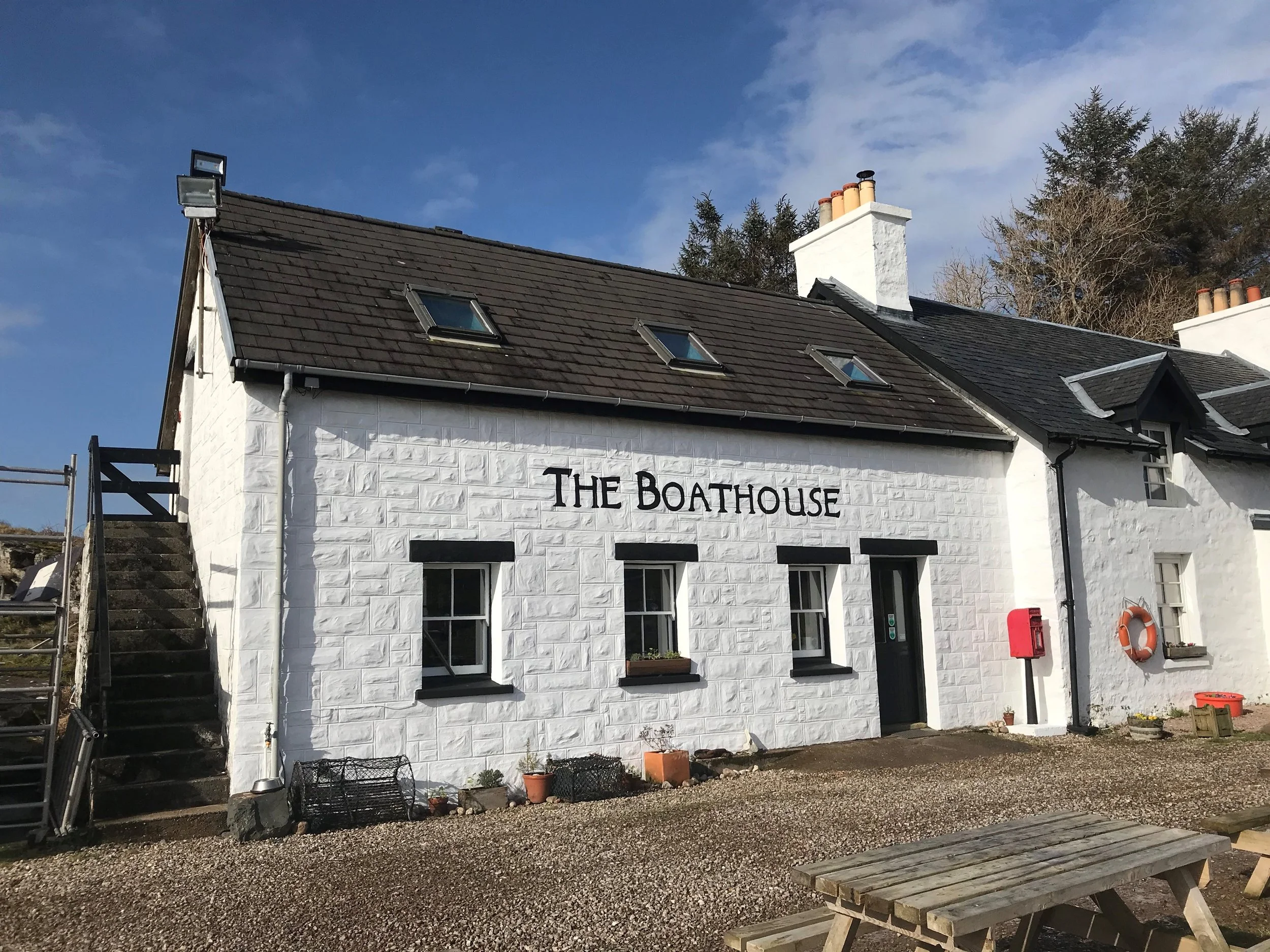 A white building with a sign that reads ��The Boathouse�� in black letters painted on the wall. The building has a dark tiled roof with three skylights and a chimney. There are three windows with black frames and flower boxes on the wall. An outdoor staircase is visible on the left side. The ground is gravel, with a wooden picnic table in the foreground and some plants and outdoor equipment around the building.