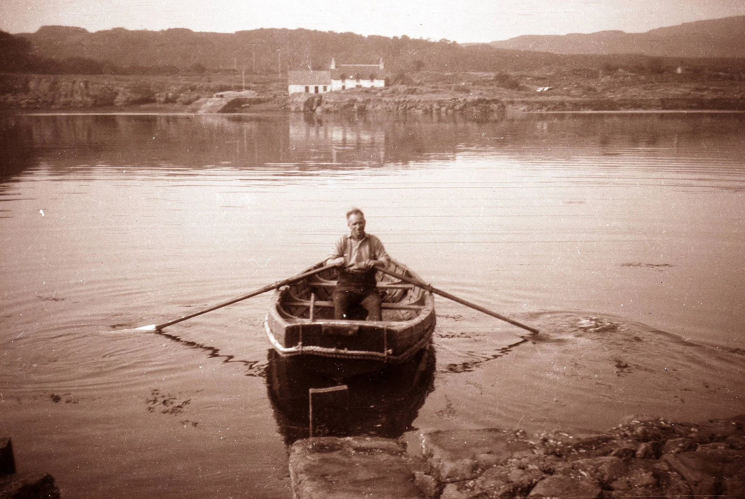 A man rowing a boat on a lake with hills and a building in the background.