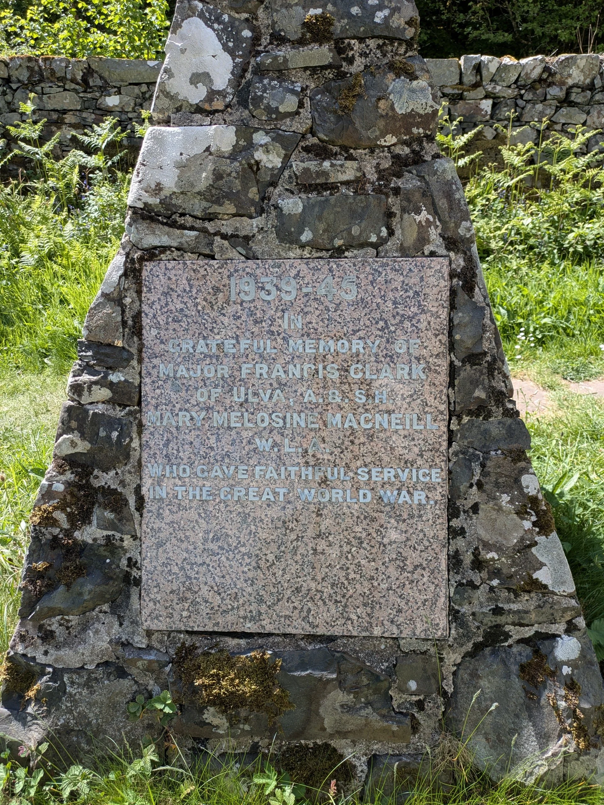 A memorial stone surrounded by grass and plants, with a plaque dedicated to Major Francis Clark and others who served in World War II, located on a field with a stone wall and greenery in the background.