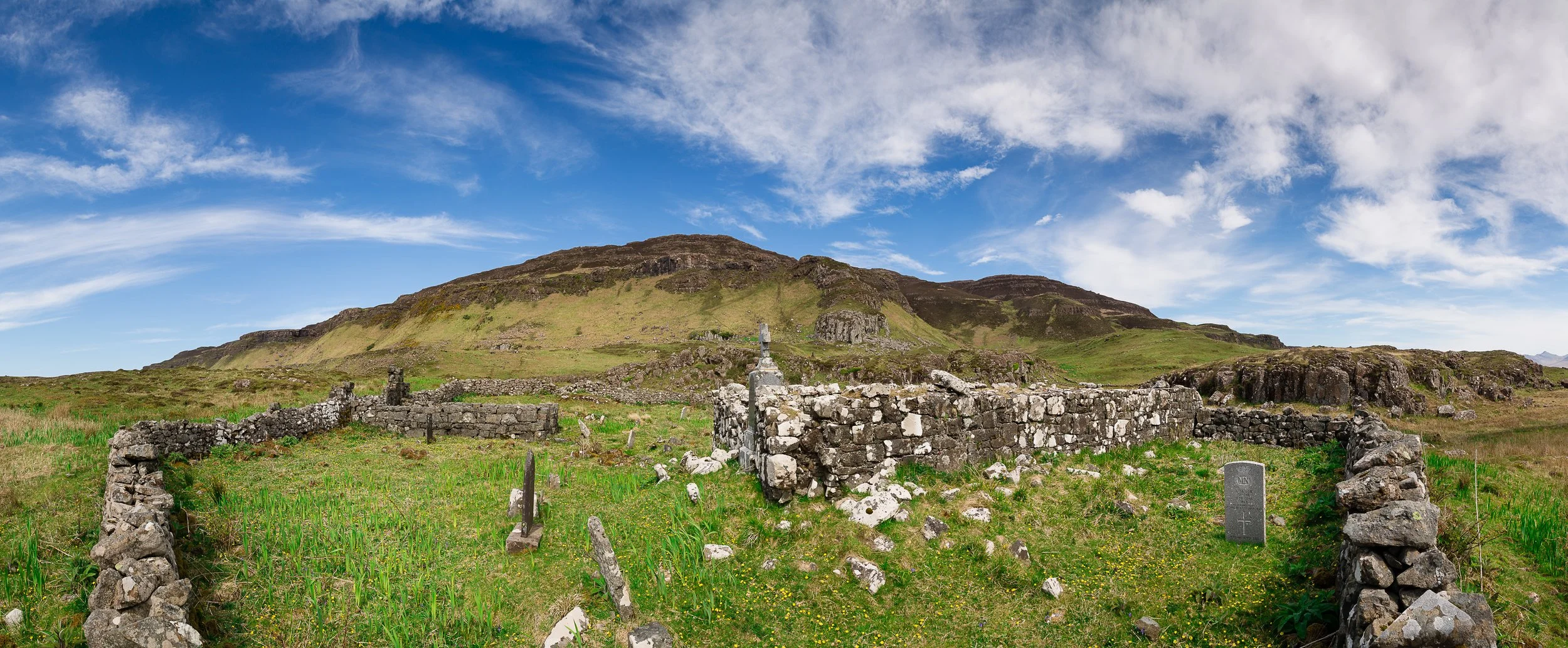 Ancient stone ruins and gravestones in a green field with a hill and blue sky with clouds in the background.