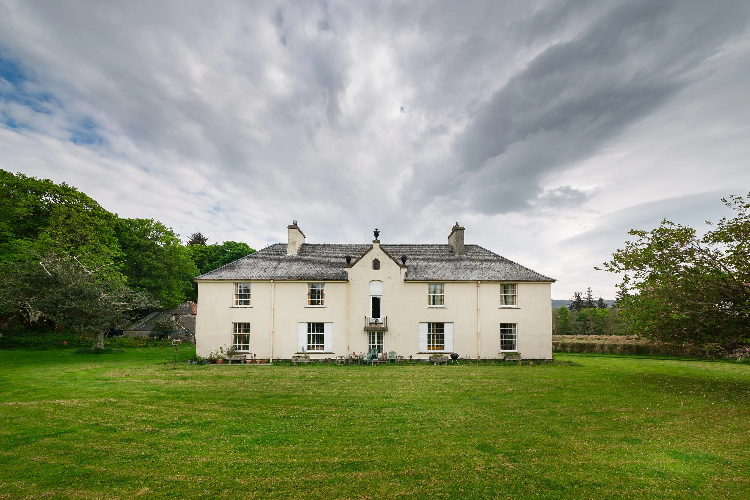 A large white house with multiple windows and a gray roof, surrounded by a green lawn and trees, under a cloudy sky.
