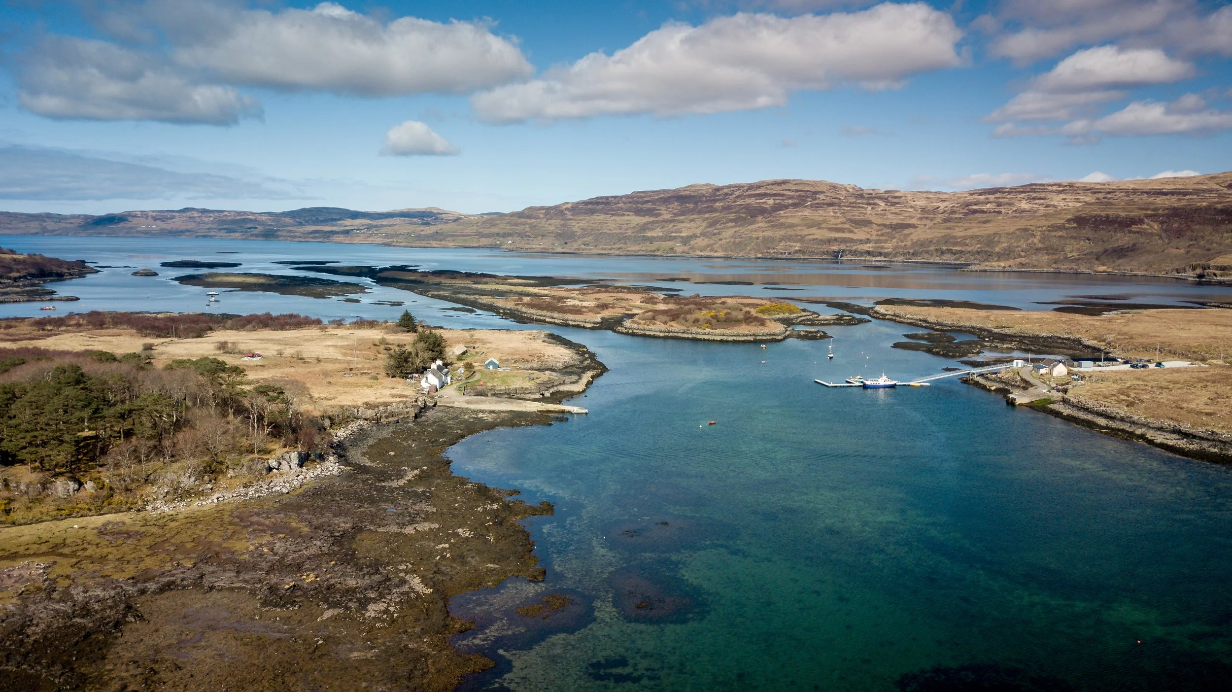A scenic aerial view of a river with several small boats, surrounding buildings, green trees, and expansive wetlands, with rolling hills and a partly cloudy sky in the background.