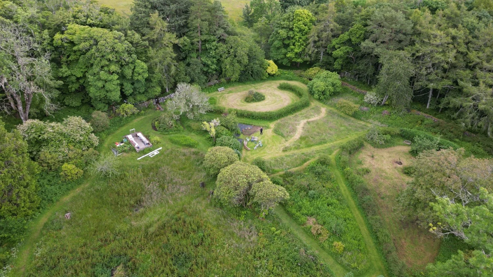 A lush green outdoor garden with various trees, shrubs, and pathways. There is a circular grassy area in the center, benches, and a person standing near some chairs. The scene is surrounded by dense forest.