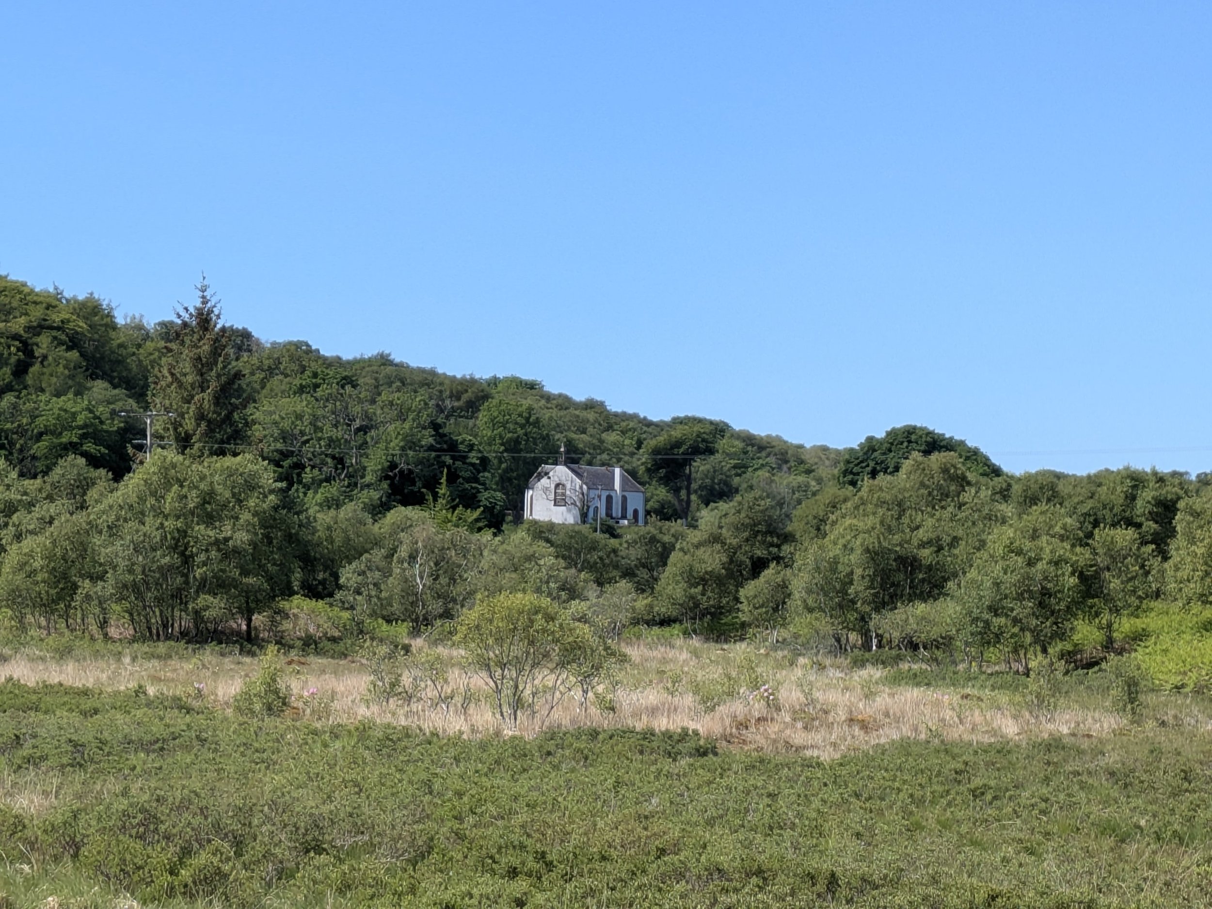 A small white church on a hill surrounded by green trees and grass under a clear blue sky.