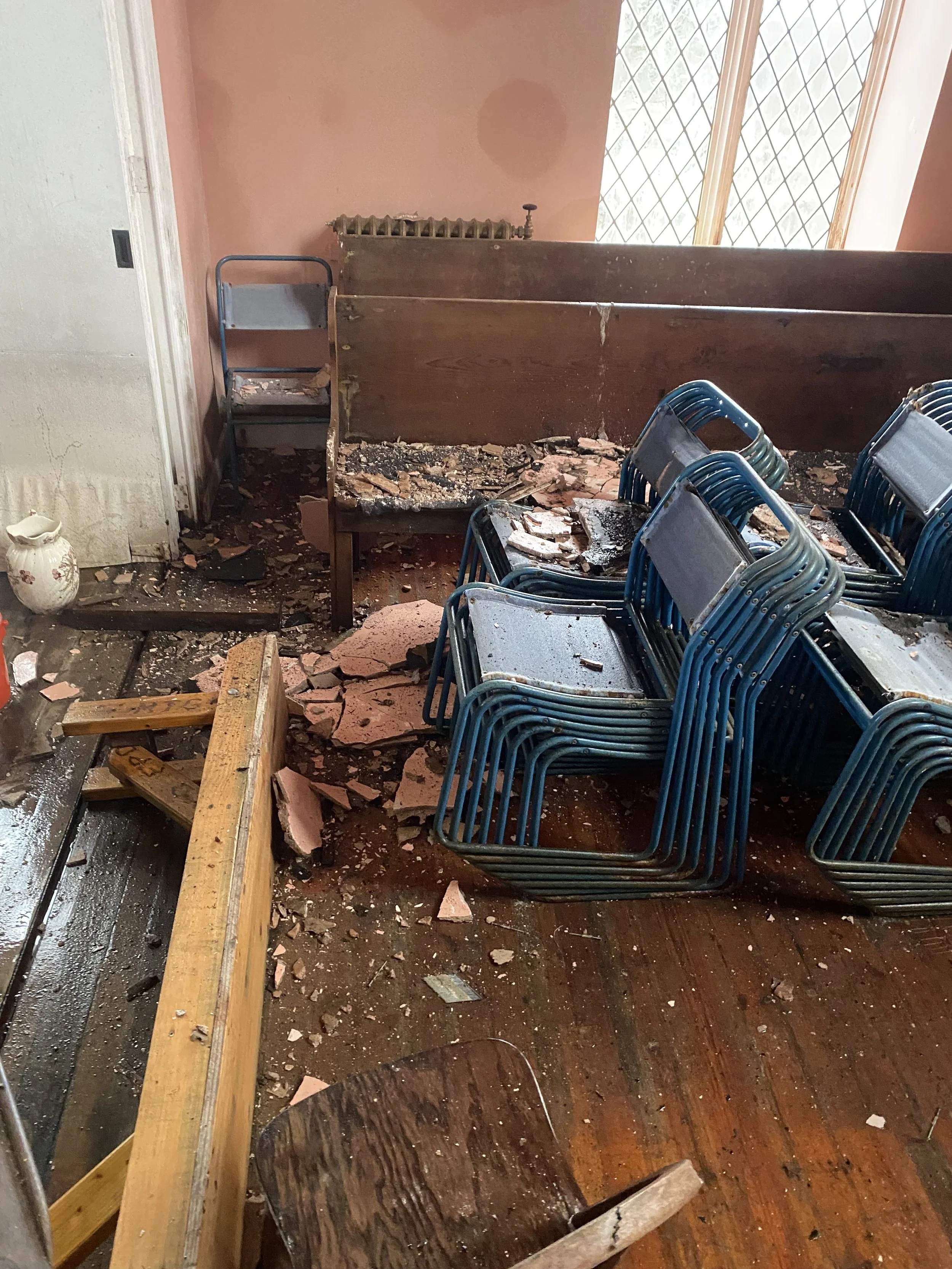 A room with overturned chairs, debris, and damage from a possible disaster, with a pink wall, radiator, and window in the background.