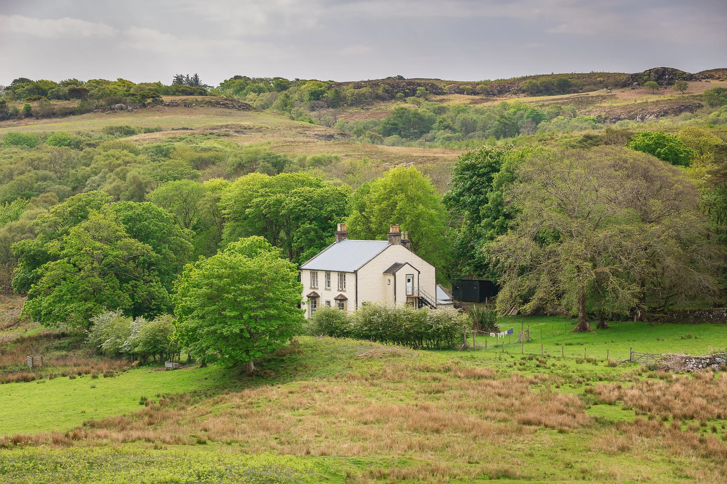 A white house with a metal roof nestled among green trees in a rural landscape, with hills in the background and a cloudy sky above.