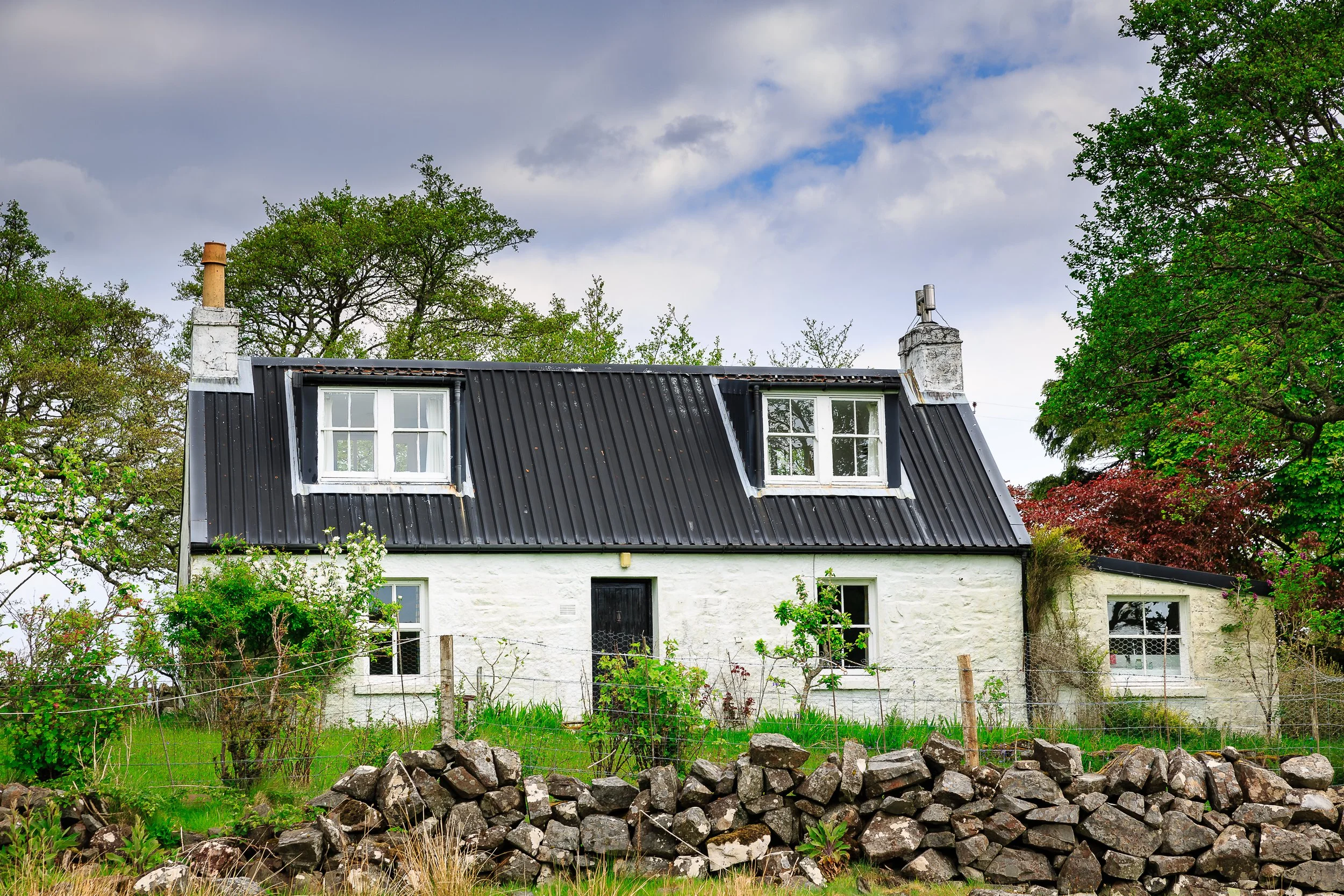 A white cottage with a black metal roof and four windows, surrounded by trees and a stone wall in the foreground.