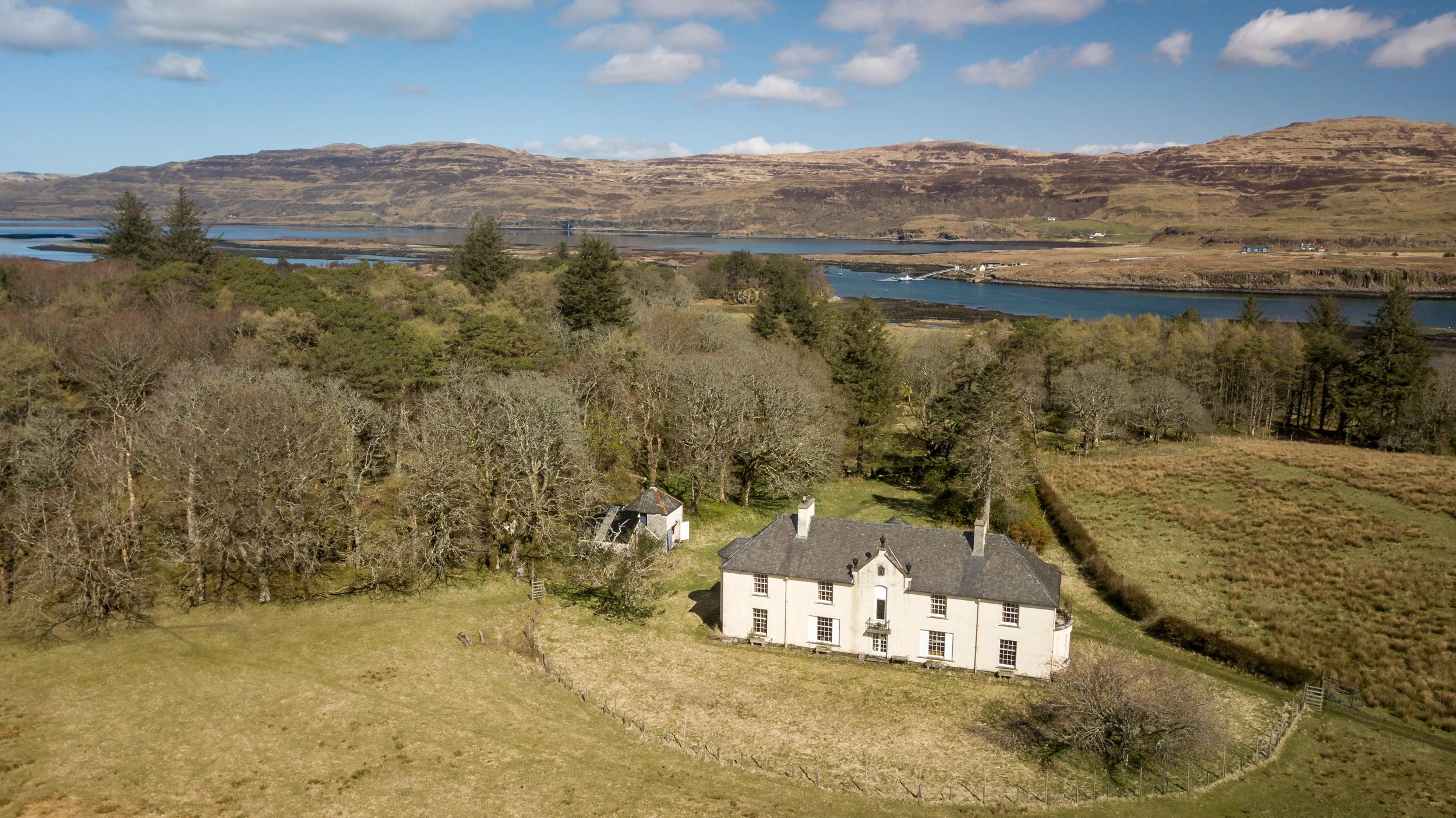 A large white historic house with multiple windows, surrounded by trees and open fields, with a river and distant hills in the background under a partly cloudy sky.