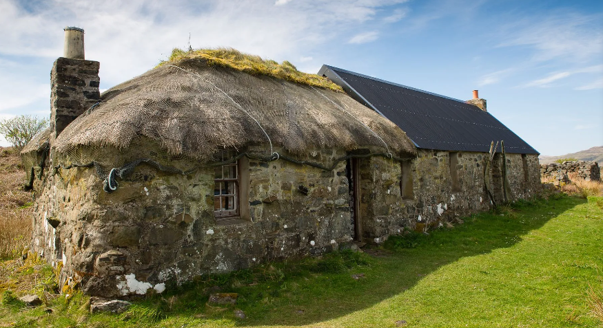 A stone cottage with a thatched roof on the left side and a metal roof on the right side, situated in a grassy landscape under a partly cloudy sky.
