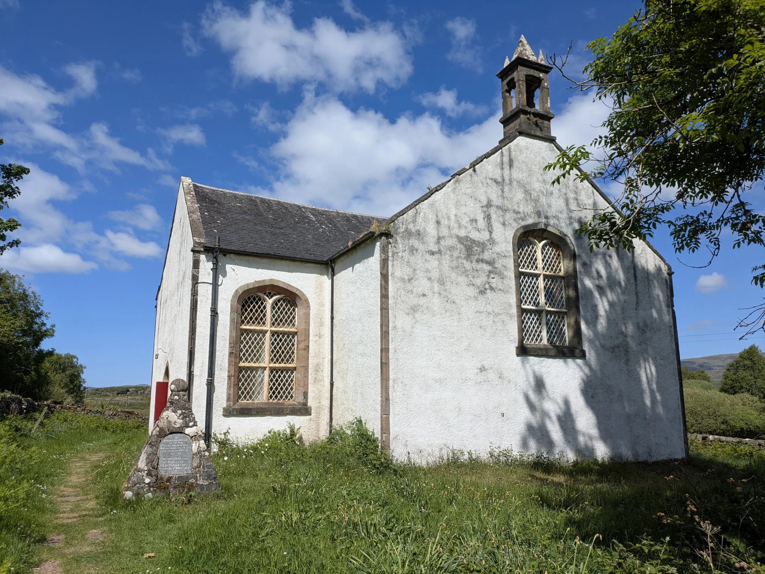 A white church with stone window frames and a small bell tower, surrounded by green grass, trees, and a partly cloudy sky.