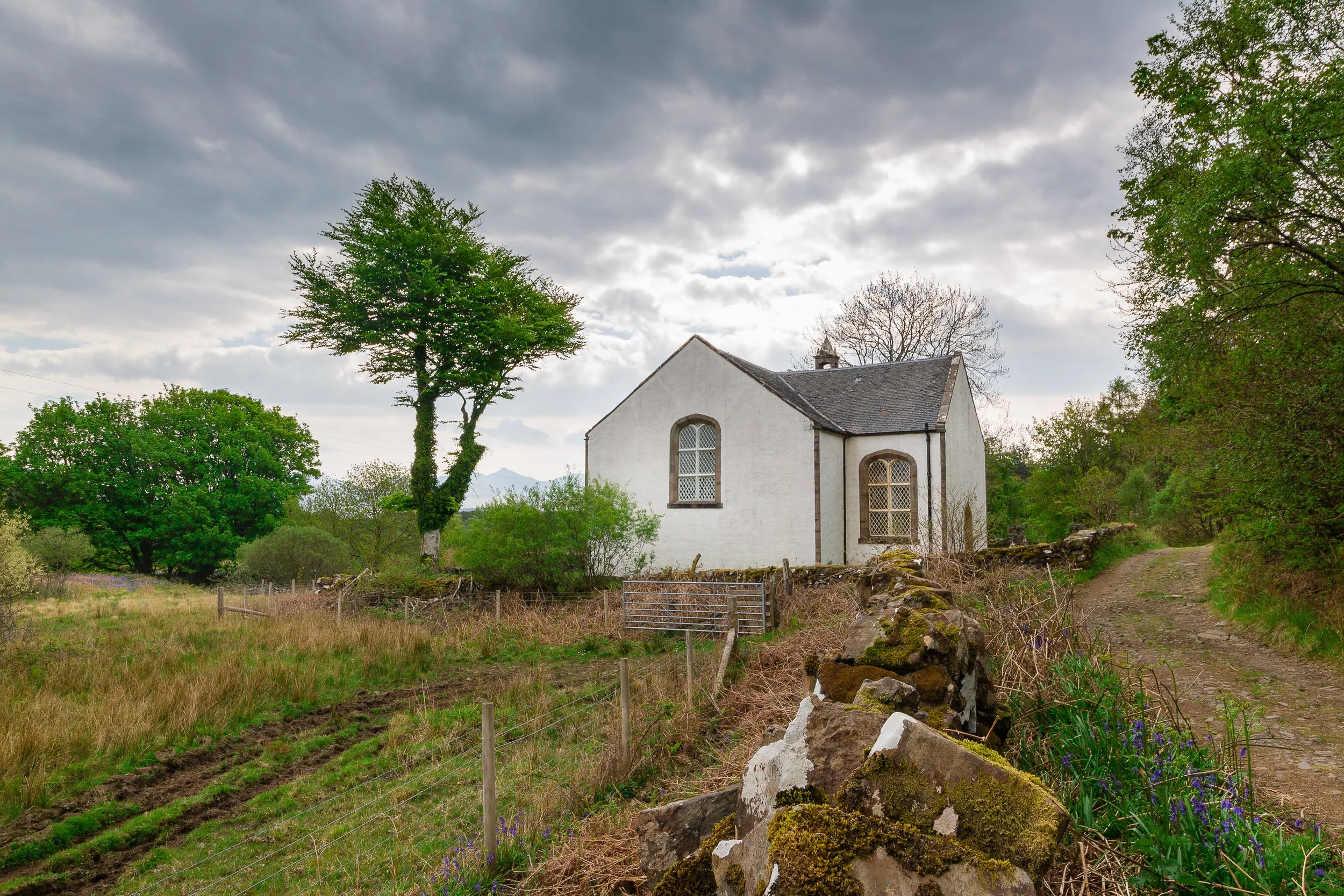 A small white church with arched windows surrounded by trees and a stone wall, with a dirt road to the right.