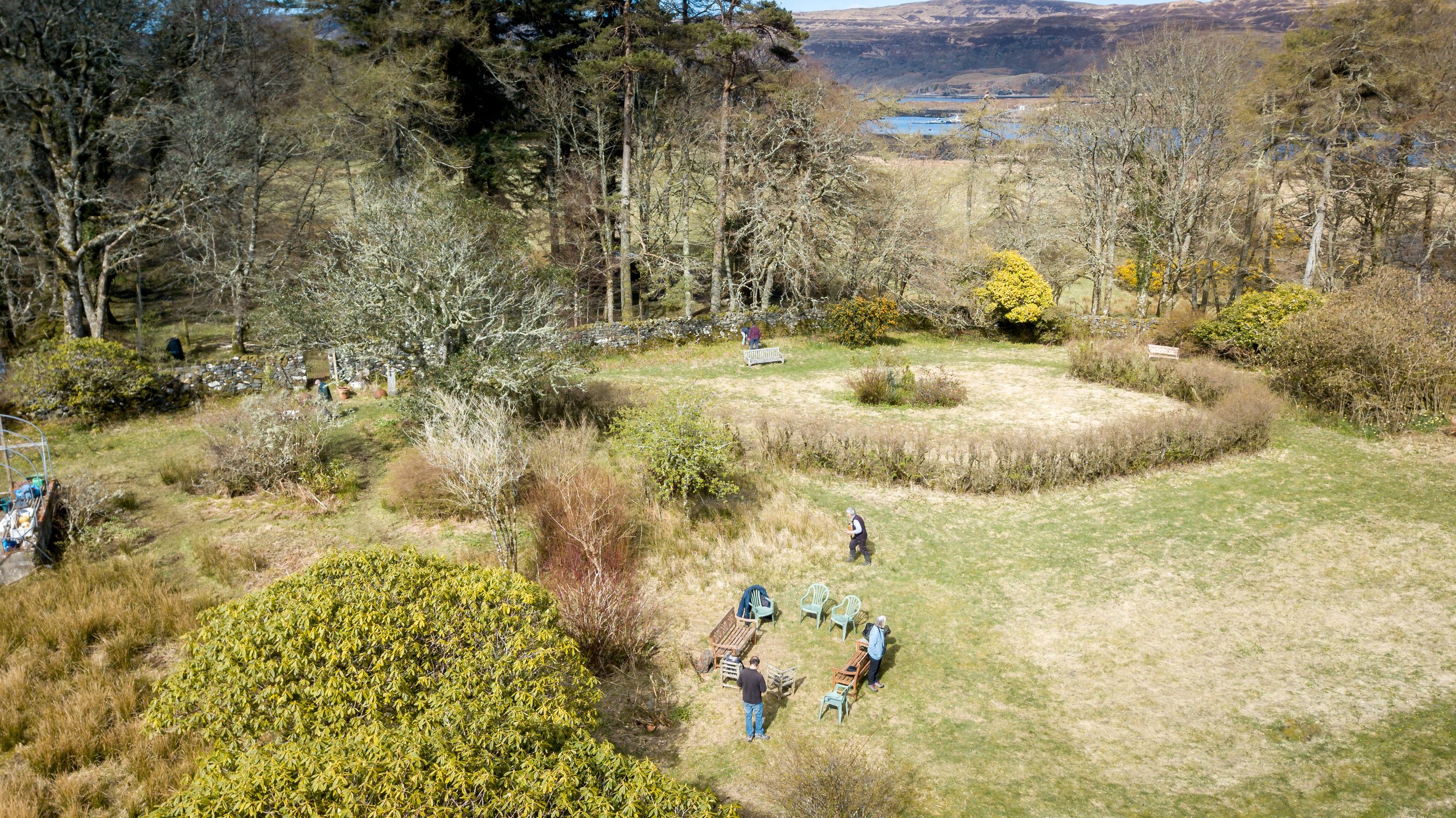 A panoramic view of a garden with trees, bushes, and open grassy areas. Several people are scattered throughout, some sitting on chairs, others standing or walking around. There are garden benches, a greenhouse, and a stone wall in the background with large trees behind it. The setting appears to be in a rural or countryside area with hills and water in the distance.