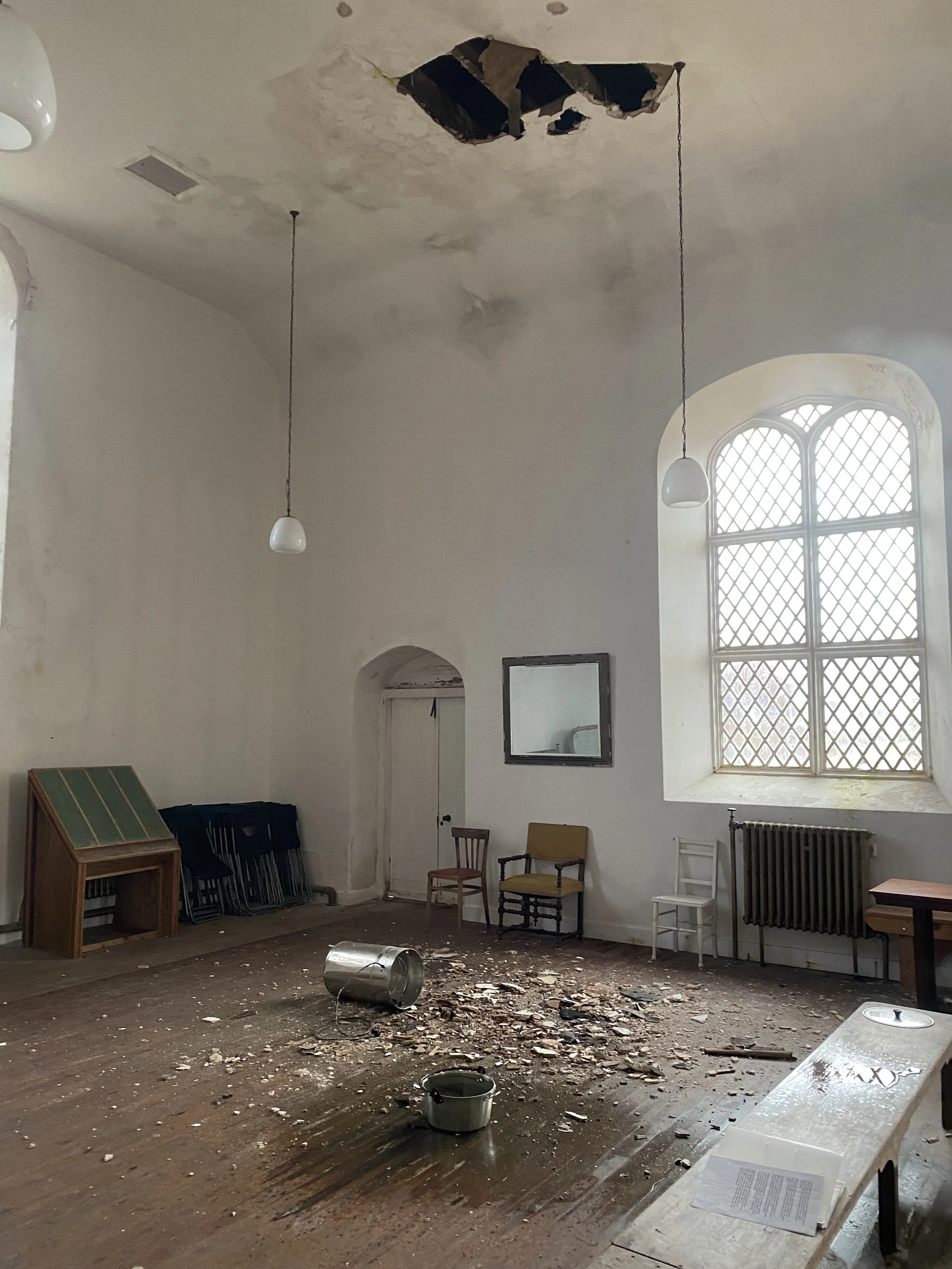 An abandoned room with peeling ceiling paint and a large hole in the ceiling, scattered debris and paint chips on the wooden floor, a tipped-over metal bucket, and several chairs against the wall near a large arched window.