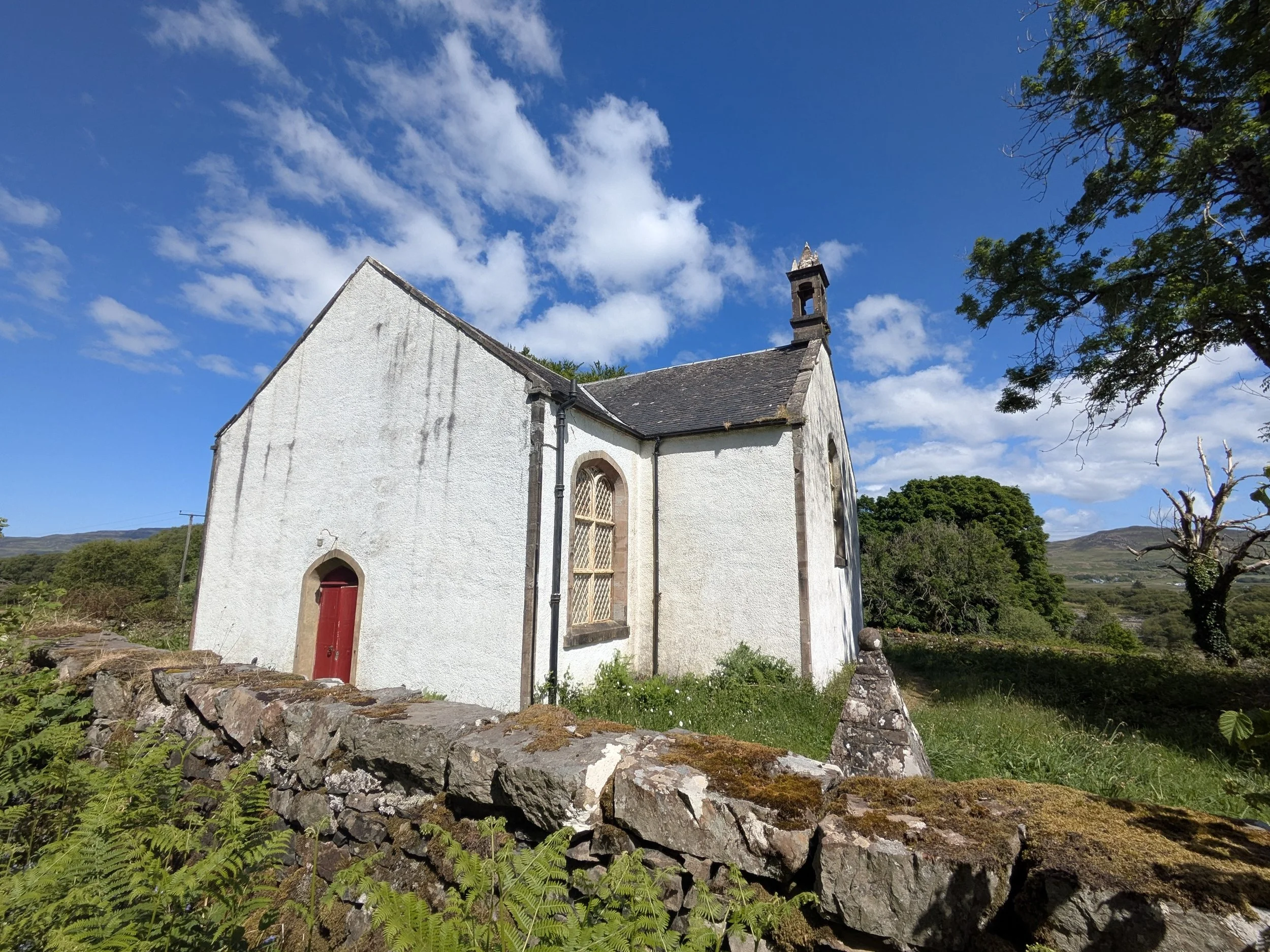 White stone church with a red door, surrounded by a stone wall, green grass, and trees, under a blue sky with clouds.