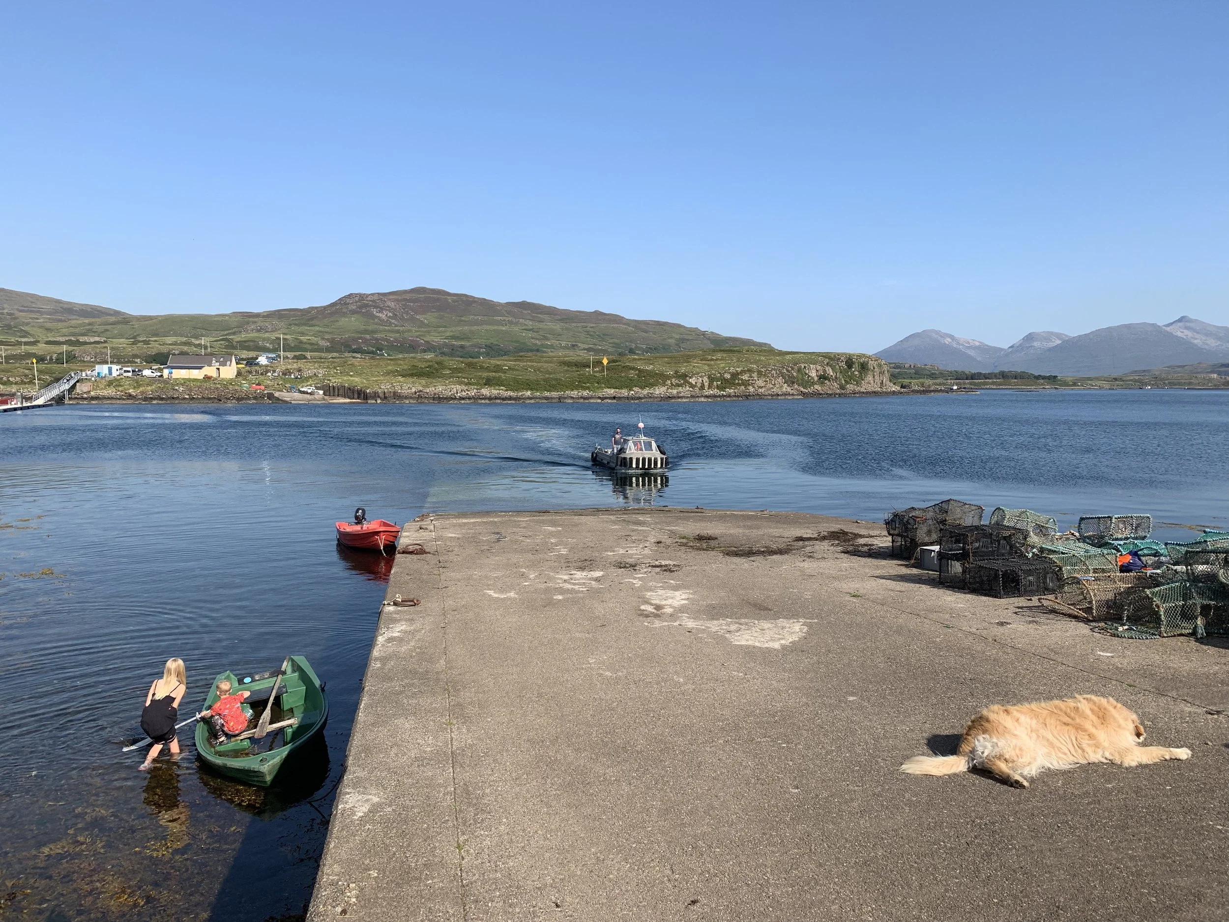 A waterfront scene with a large concrete dock, two small boats (one green with a woman and child, and one red), a motorboat in the water, lobster traps on the dock, a dog lying on the dock, and hills with mountains in the background under a clear blue sky.