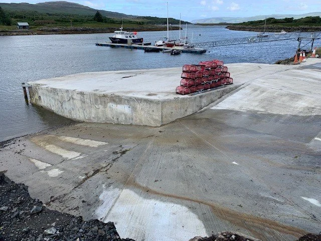 Concrete dock with a stack of red buoys, boats anchored nearby, and a hilly landscape in the background.