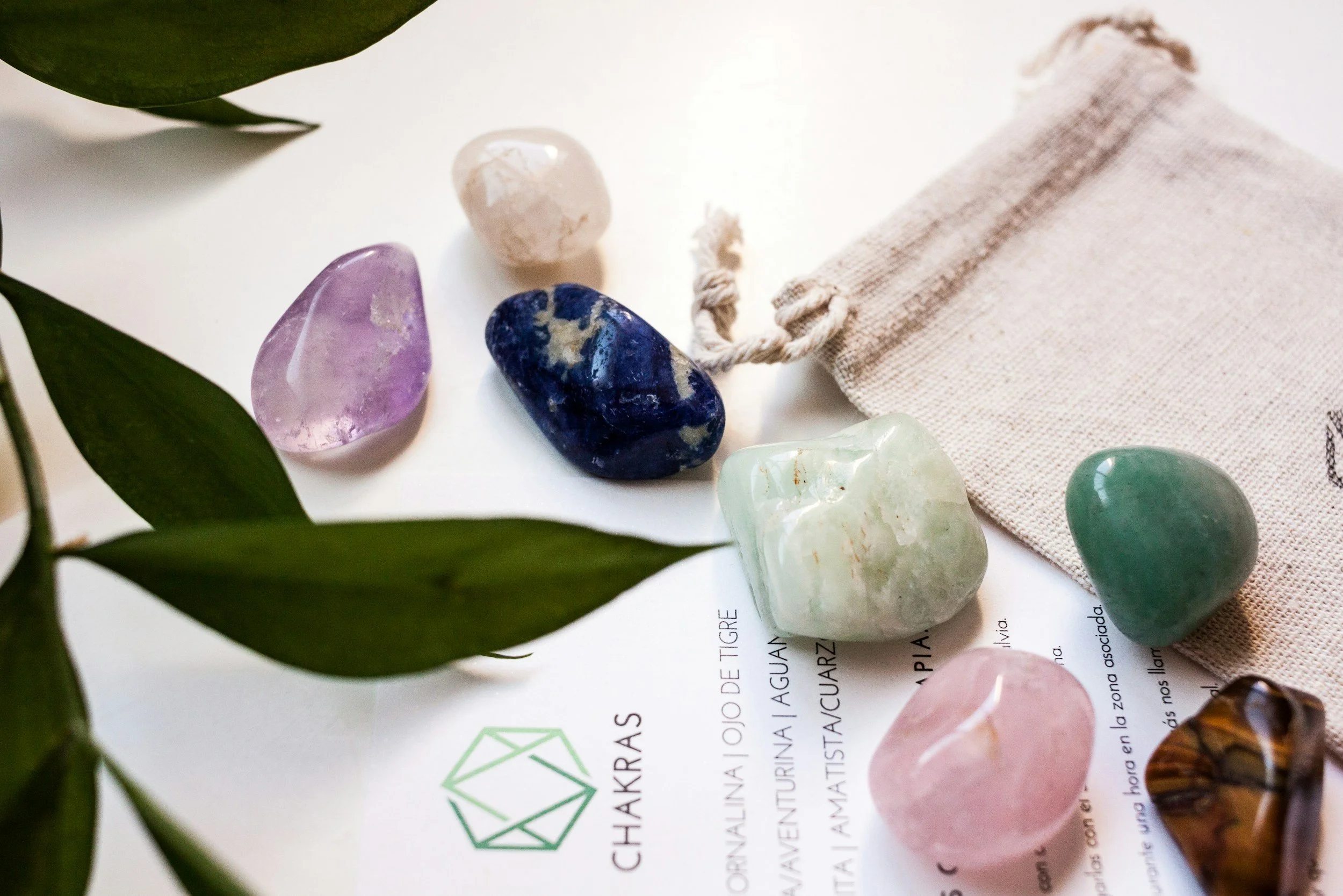 Assorted polished gemstones including amethyst, lapis lazuli, aventurine, rose quartz, and sunstone, placed on a white surface with a beige cloth bag and green leaves nearby.