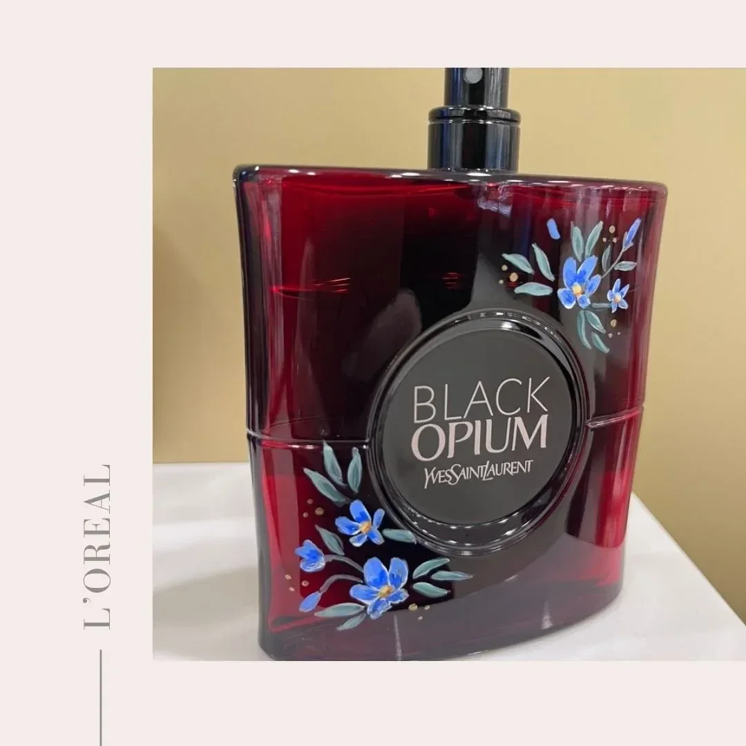 A deep ruby red BLACK OPIUM YVES SAINT LAURENT fragrance bottle with a dark glossy finish and black spray nozzle, customised with hand-painted blue flowers, green leaves with gold dot details on the front of the bottle.