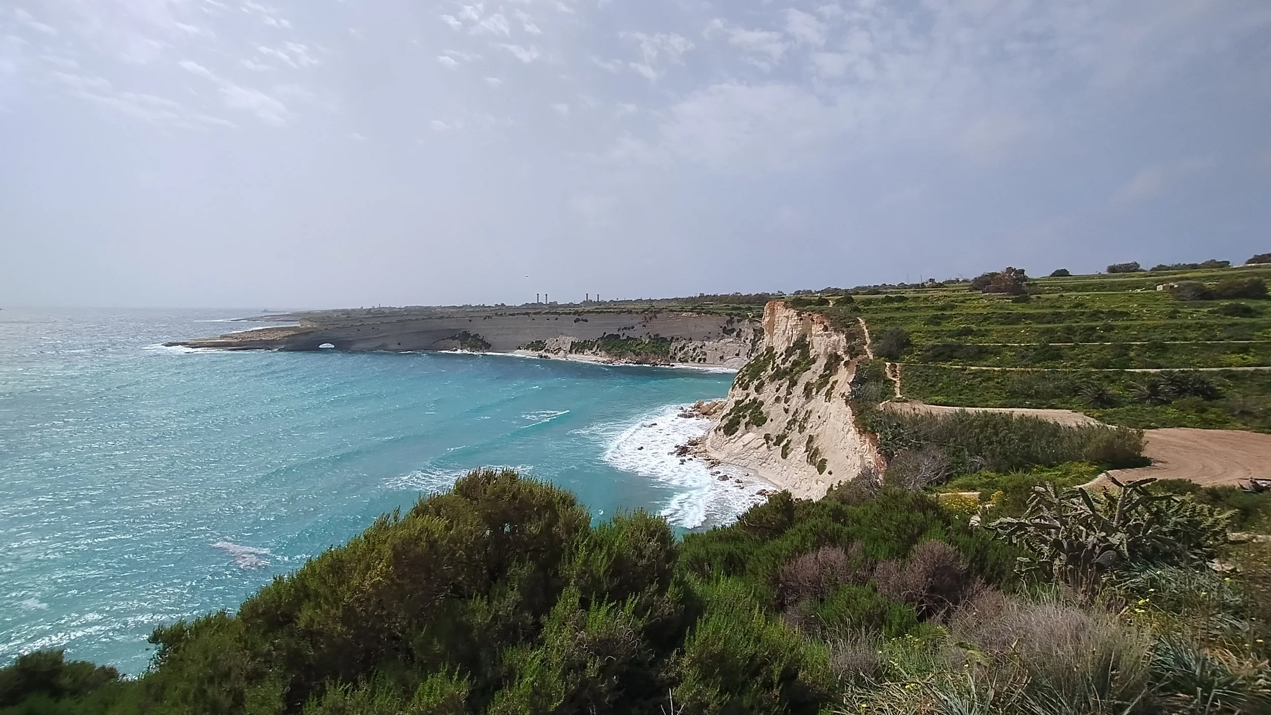 Coastal cliffs and green hills overlooking a turquoise ocean with a cloudy sky.
