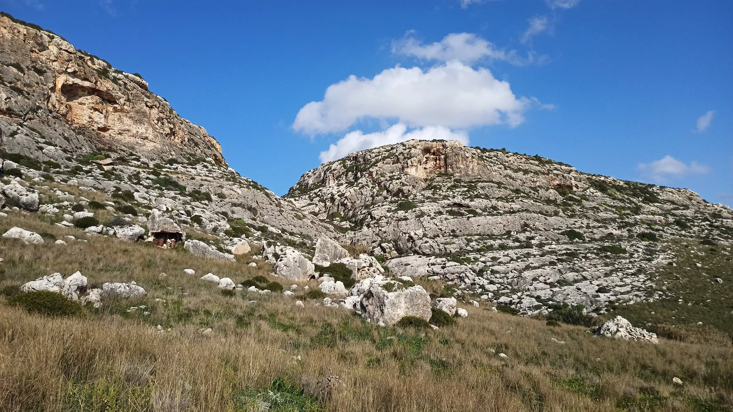 A mountain landscape with rocky cliffs, a blue sky with some white clouds, and grassy terrain with scattered rocks and small shrubs.